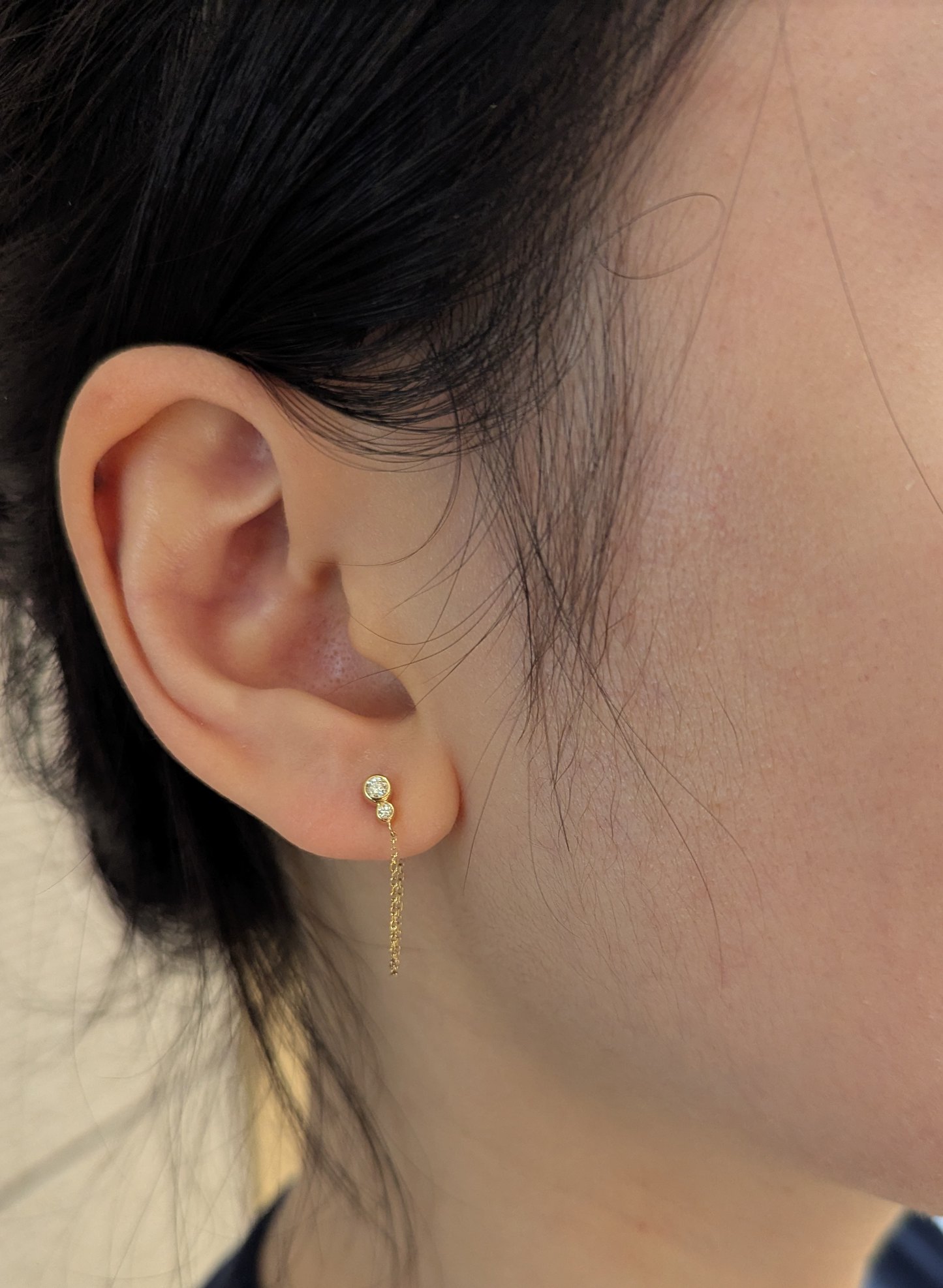 Close-up on ear showing a 10K yellow gold 0.07 cttw Canadian diamond stud earring with two bezel-set diamonds and a delicate dangling chain.