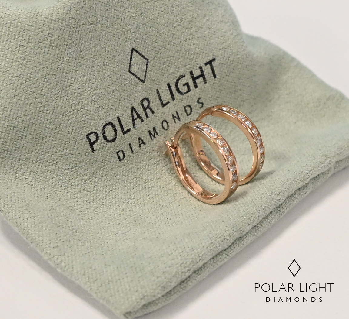 A pair of 10K rose gold huggie hoop earrings, featuring channel-set Canadian diamonds, displayed on top of a soft, iridescent "polar light" jewelry pouch that subtly shows the brand logo.