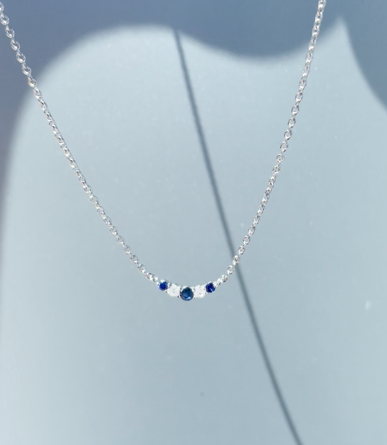 Close-up product view of a 925 sterling silver V-shaped curve necklace suspended against a grey background, showcasing a 3mm curved row of alternating round-cut blue sapphires and brilliant Canadian diamonds.