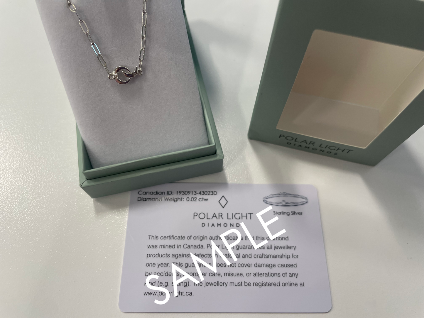A product presentation and authenticity display for a Polar Light Diamonds celestial bracelet. A sterling silver paperclip chain bracelet featuring an interlocking crescent moon and circle motif with a 0.02 cttw Canadian diamond accent is showcased inside an open, sage green gift box. The branded outer gift box lid sits alongside it, positioned directly above a matching white Polar Light Diamonds certificate of origin sample validating its ethical Canadian mining background and quality specifications.