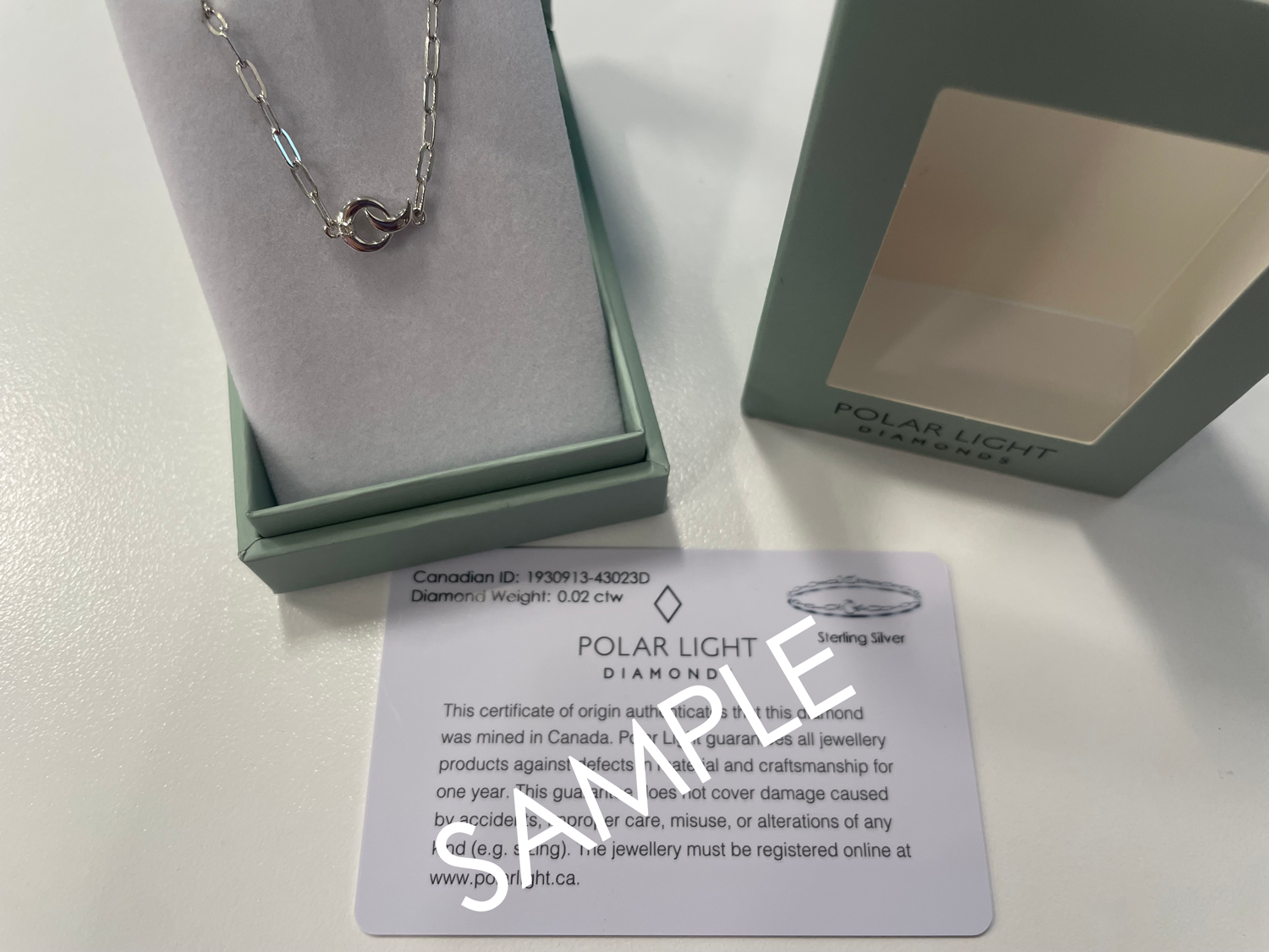 A product presentation and authenticity display for a Polar Light Diamonds celestial bracelet. A sterling silver paperclip chain bracelet featuring an interlocking crescent moon and circle motif with a 0.02 cttw Canadian diamond accent is showcased inside an open, sage green gift box. The branded outer gift box lid sits alongside it, positioned directly above a matching white Polar Light Diamonds certificate of origin sample validating its ethical Canadian mining background and quality specifications.