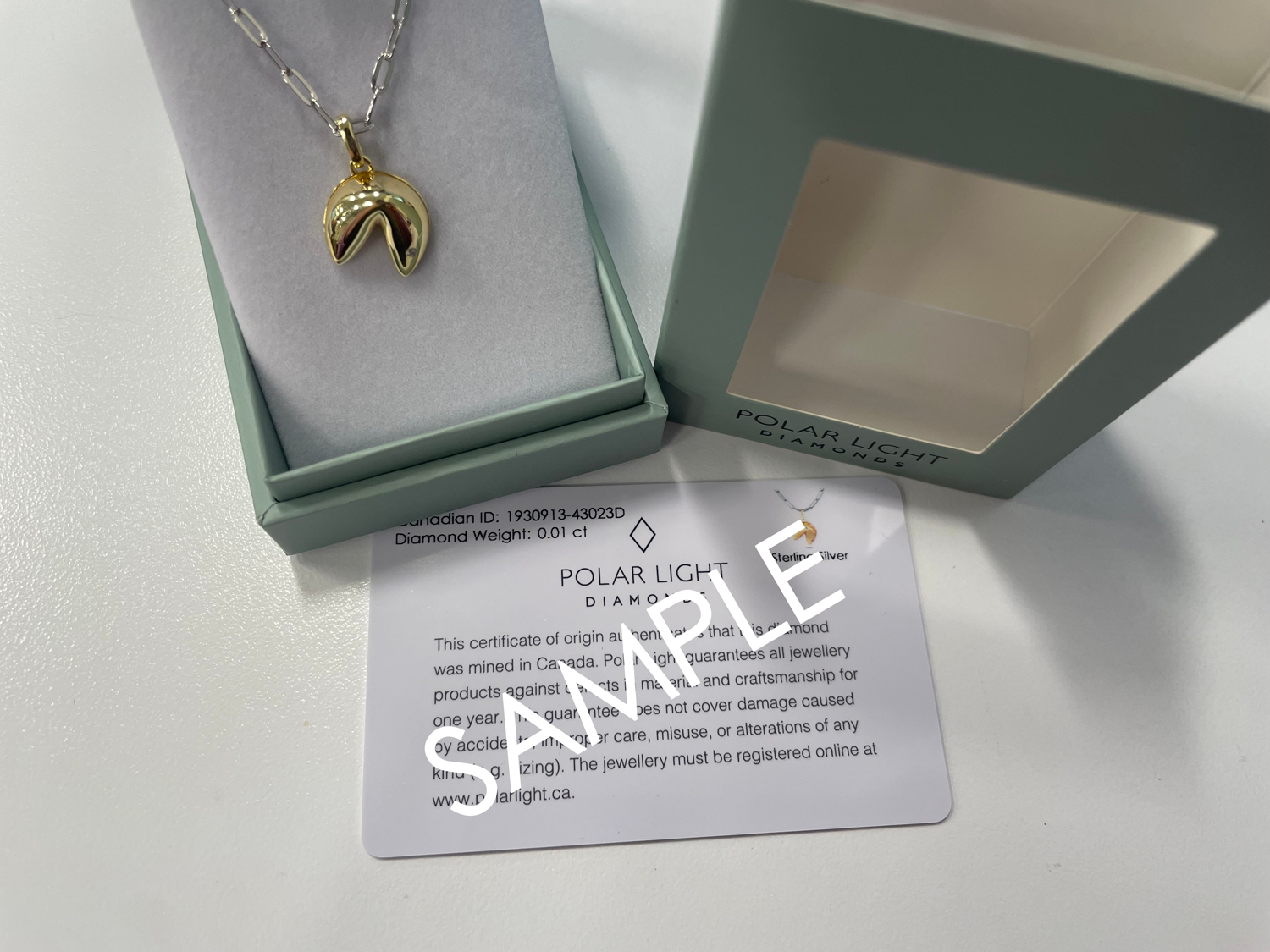 A two-tone fortune cookie pendant necklace displayed in a sage green Polar Light Diamonds gift box next to its certificate of origin card marked "SAMPLE."