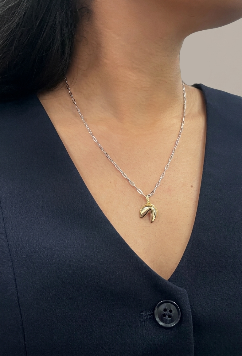 A model wearing a two-tone pendant necklace, featuring a polished gold-plated sterling silver fortune cookie charm hanging from a sterling silver paperclip link chain, showcasing how it sits on the neckline.