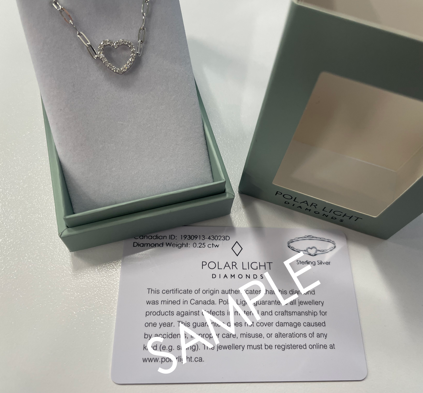 A product presentation and authenticity display for a diamond Polar Light Diamonds bracelet. A 925 sterling silver paperclip chain bracelet featuring an open heart station lined with 0.25 cttw of sparkling Canadian diamonds is beautifully presented inside an open, sage green gift box. The signature outer box lid sits alongside it displaying the brand logo, positioned above a white matching Polar Light Diamonds certificate of origin sample card verifying its ethical Canadian origin.
