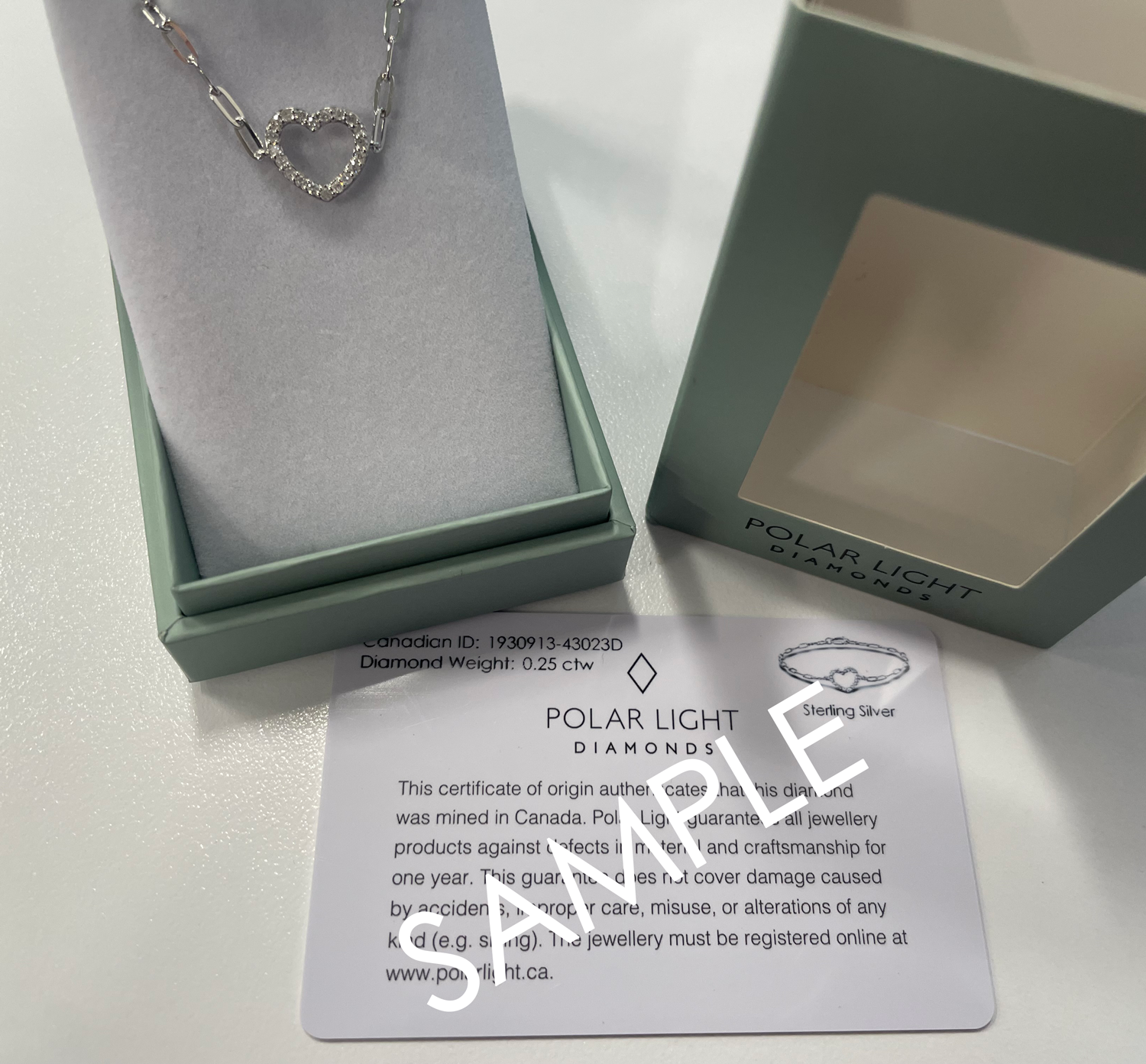 A product presentation and authenticity display for a diamond Polar Light Diamonds bracelet. A 925 sterling silver paperclip chain bracelet featuring an open heart station lined with 0.25 cttw of sparkling Canadian diamonds is beautifully presented inside an open, sage green gift box. The signature outer box lid sits alongside it displaying the brand logo, positioned above a white matching Polar Light Diamonds certificate of origin sample card verifying its ethical Canadian origin.