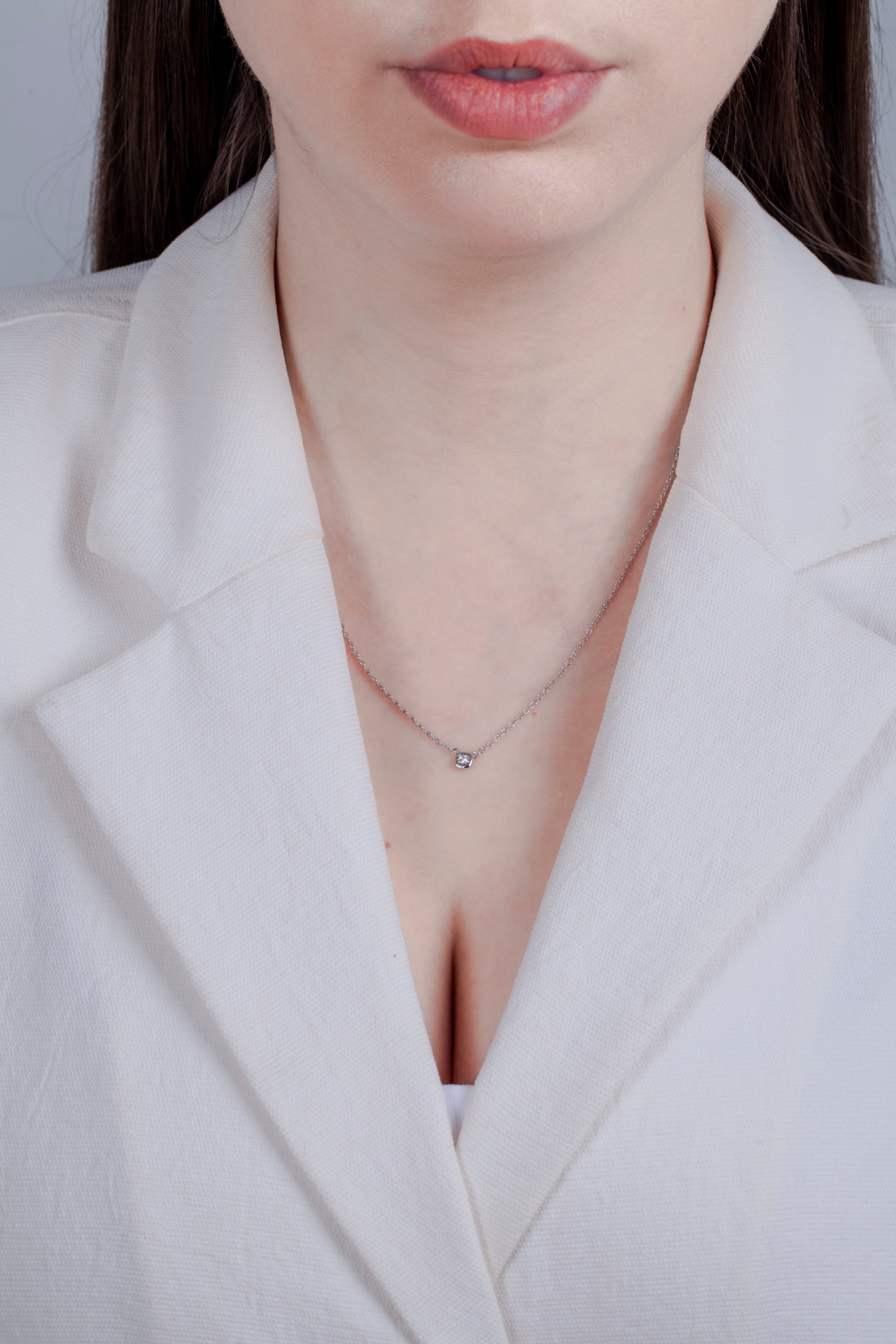 Close-up lifestyle shot of a Canadian diamond solitaire necklace on a model. The sterling silver pendant features a cushion-shaped miracle plate and bezel setting for a bold look, paired with a classic rolo chain and a professional white blazer.