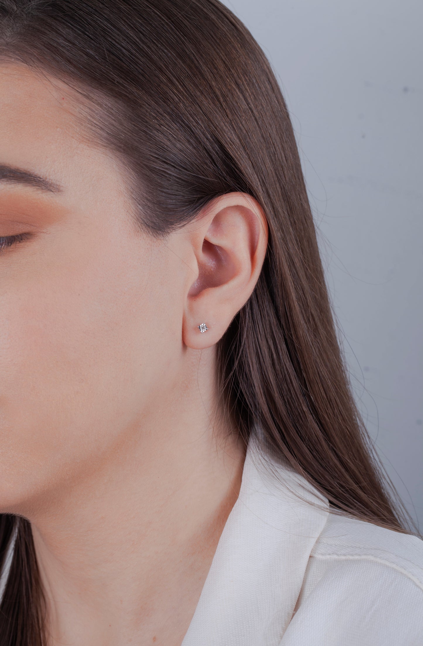 Close-up of a model with long brown hair wearing a dainty Canadian diamond stud earring. She is wearing a white blazer against a neutral background, highlighting a sophisticated, minimalist look.