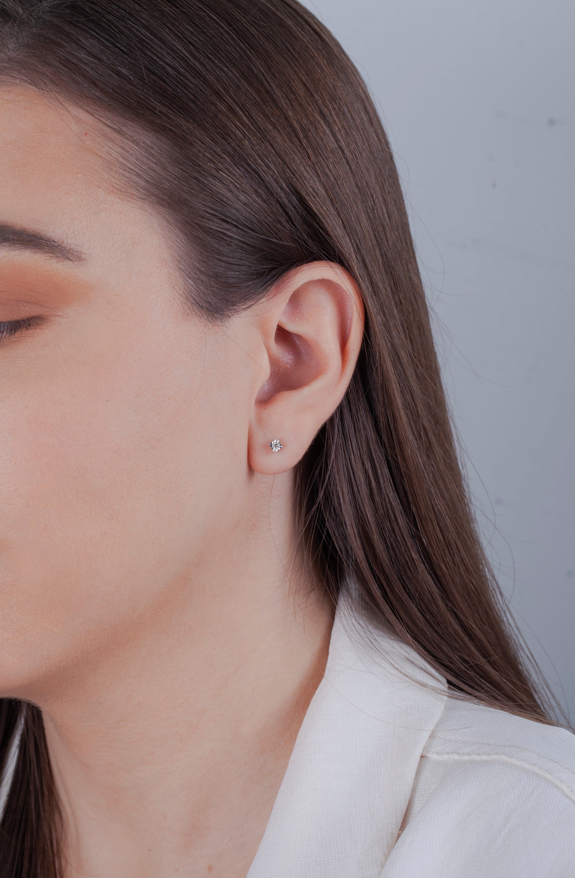 Close-up of a model with long brown hair wearing a dainty Canadian diamond stud earring. She is wearing a white blazer against a neutral background, highlighting a sophisticated, minimalist look.