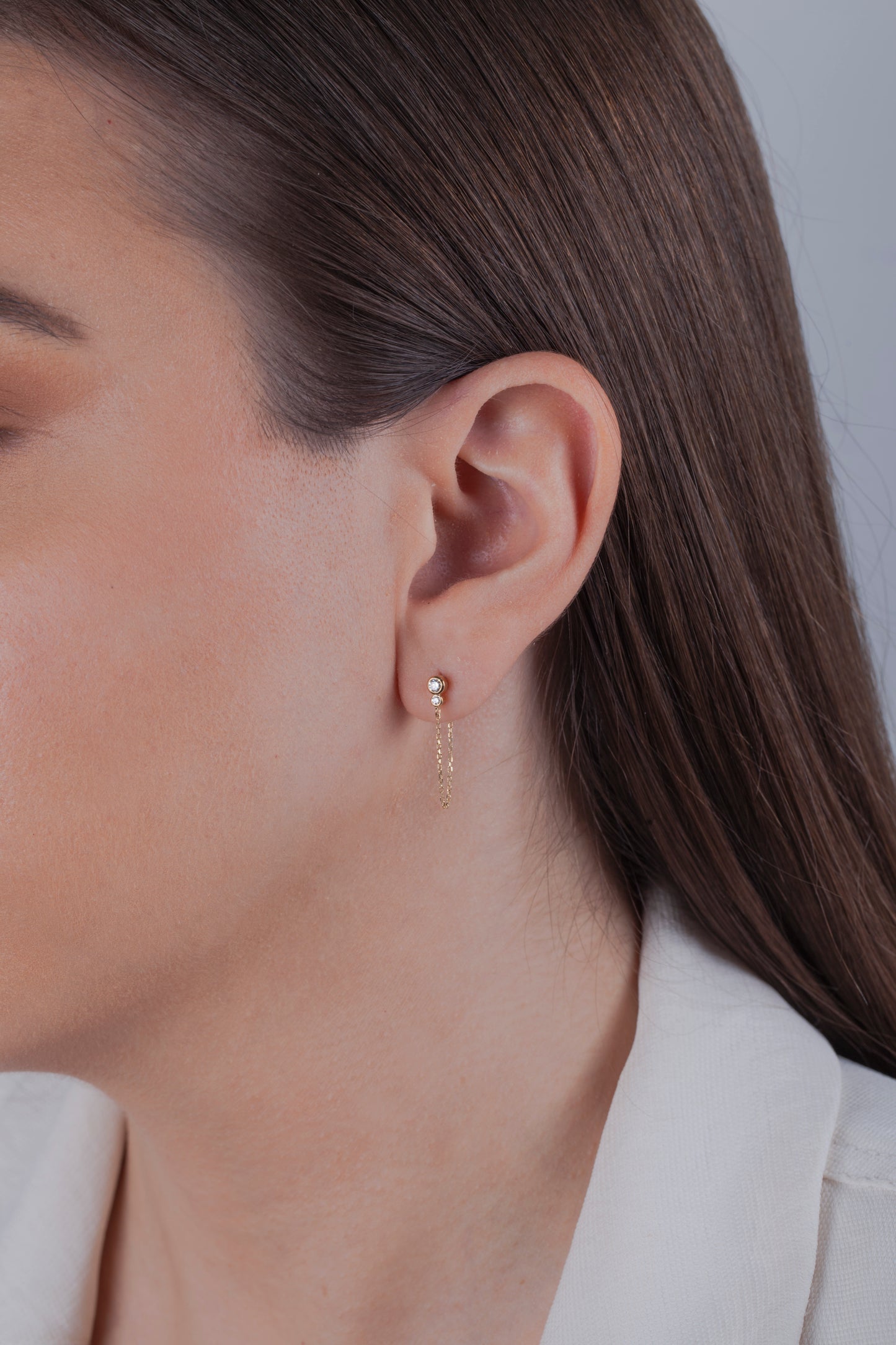 Close-up of a person wearing gold diamond stud earrings with dangle chains on a neutral background