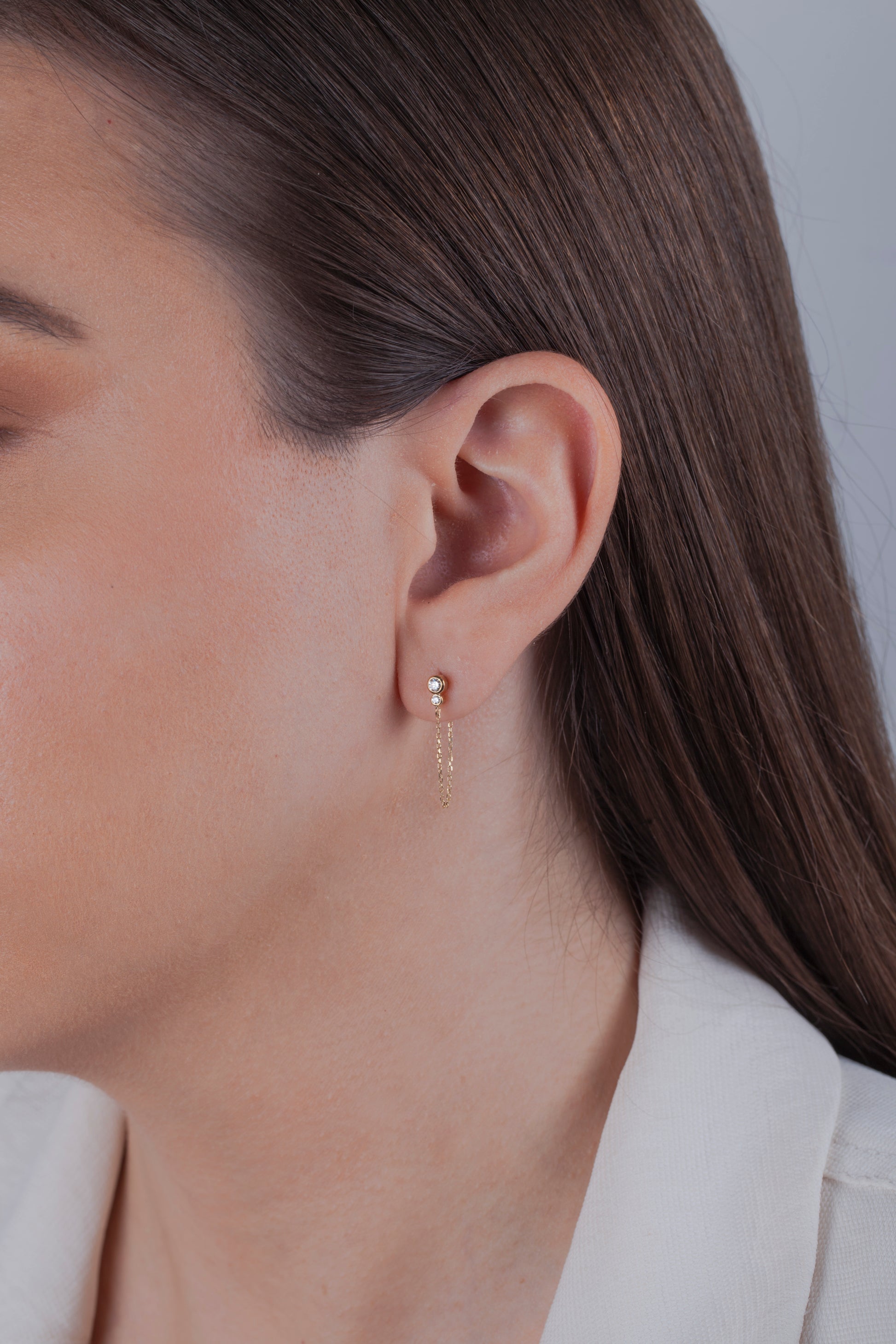 Close-up of a person wearing gold diamond stud earrings with dangle chains on a neutral background