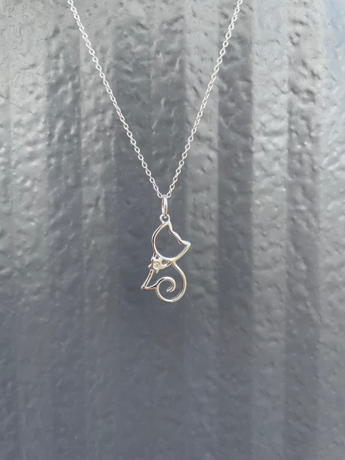 Close-up of a sterling silver kitty cat-silhouette pendant necklace, featuring a genuine Canadian diamond accent and a rolo chain against a textured grey background.
