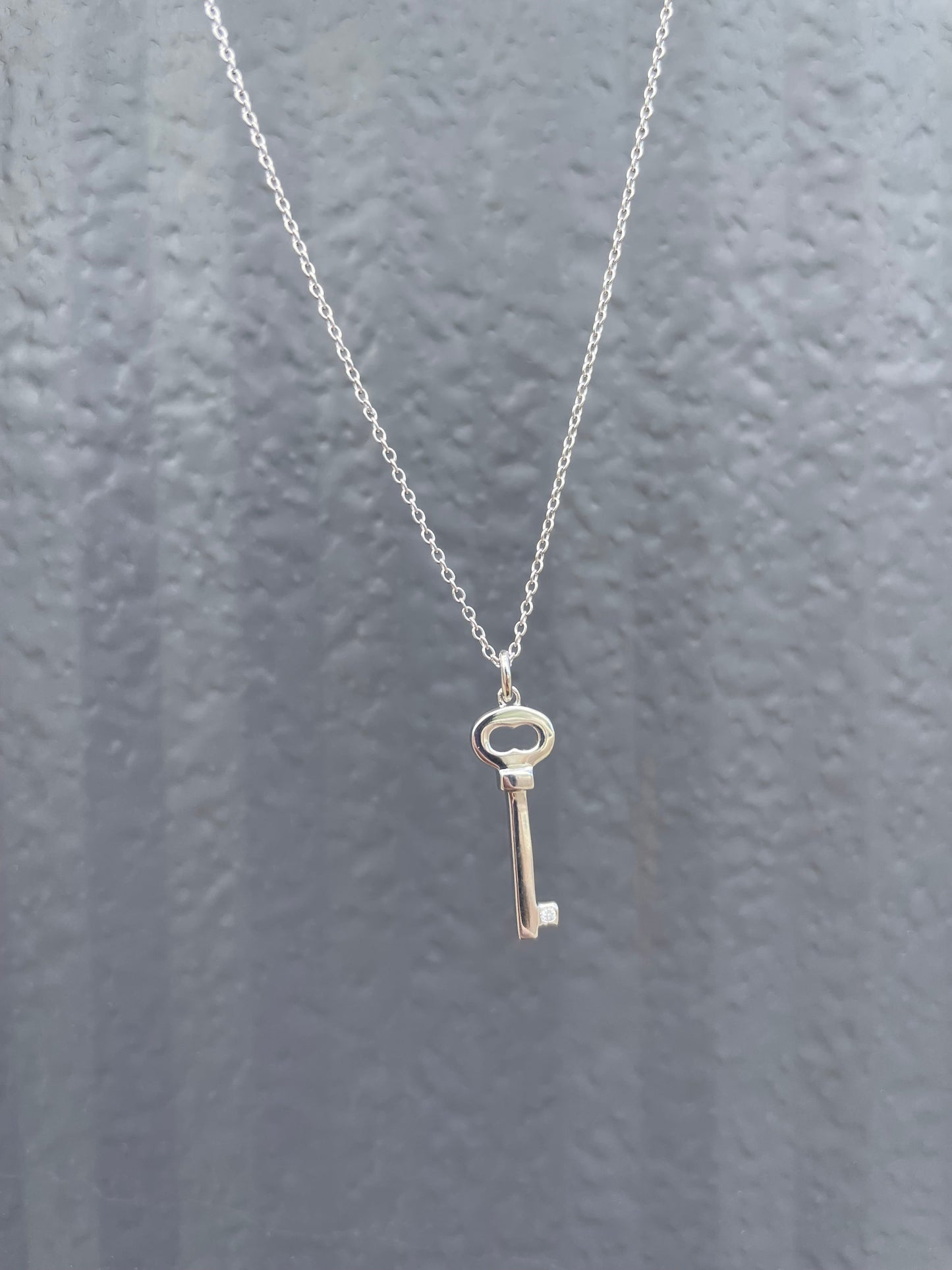 Close-up of a sterling silver key-shaped pendant, featuring a subtle Canadian diamond accent and a delicate cable chain against a textured grey background.