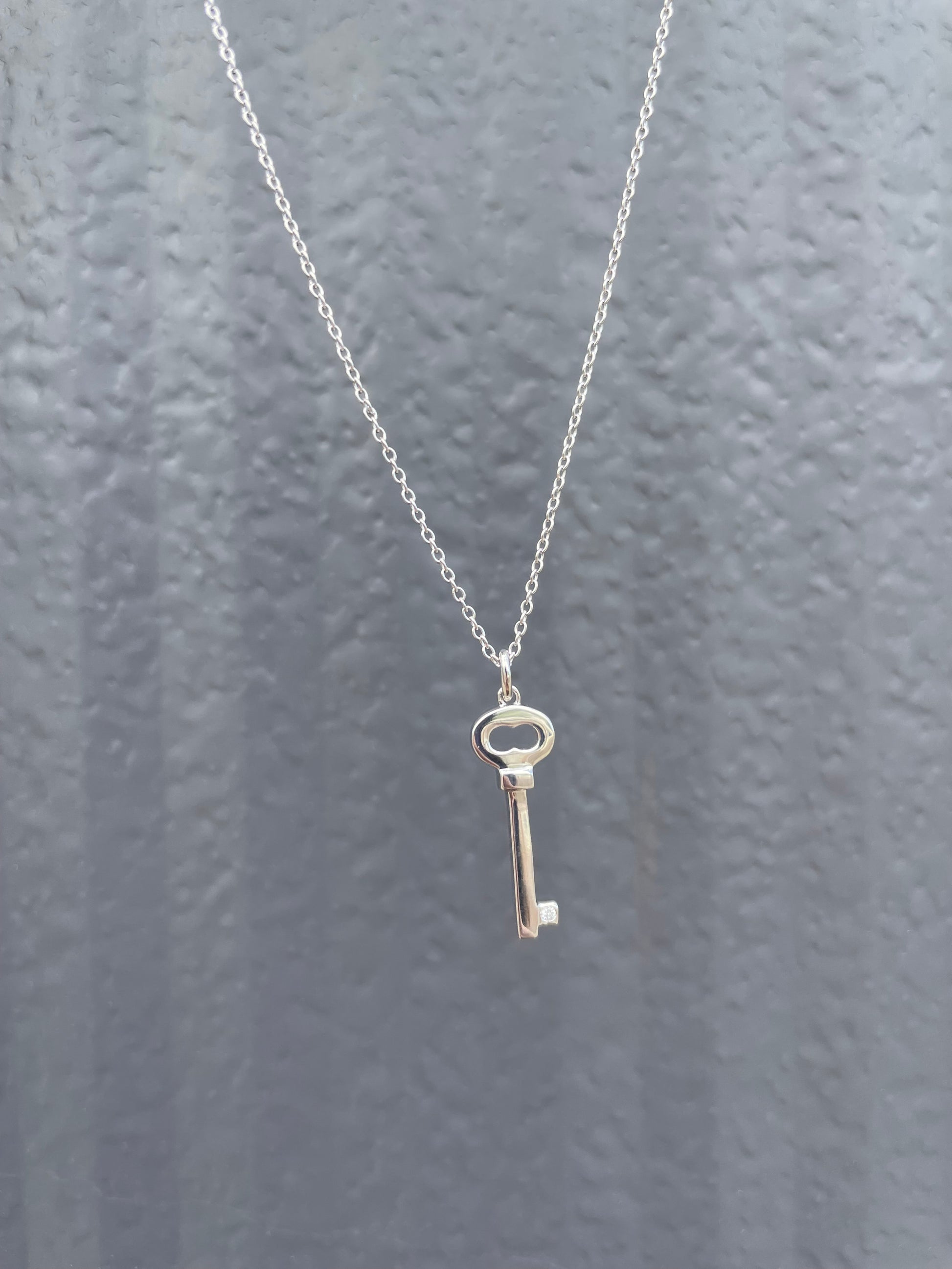 Close-up of a sterling silver key-shaped pendant, featuring a subtle Canadian diamond accent and a delicate cable chain against a textured grey background.