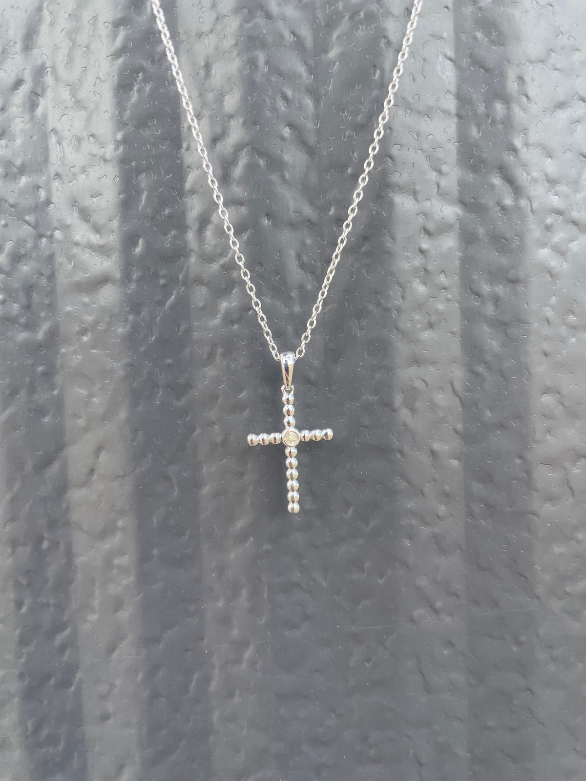 Close-up of a sterling silver beaded cross pendant, featuring a central Canadian diamond accent and a delicate rolo chain against a textured grey background.