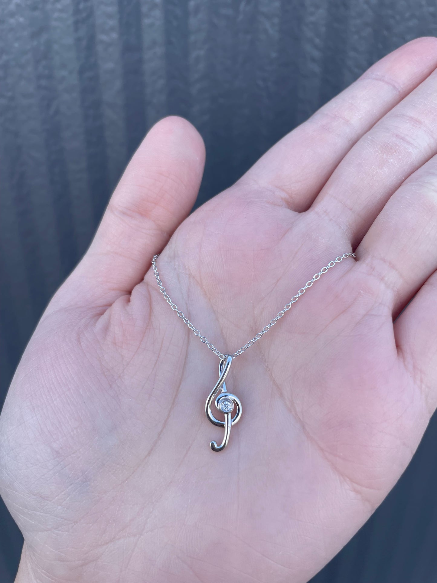 Hand holding a sterling silver treble clef pendant necklace with a genuine Canadian diamond, featuring a delicate rolo chain design.