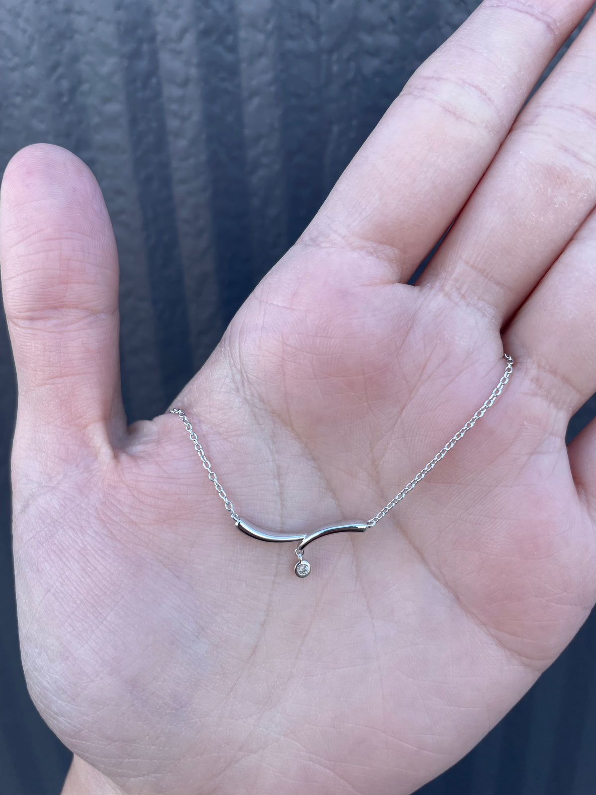 Hand holding a sterling silver curved bar necklace with a single bezel-set Canadian diamond accent, featuring a delicate chain.
