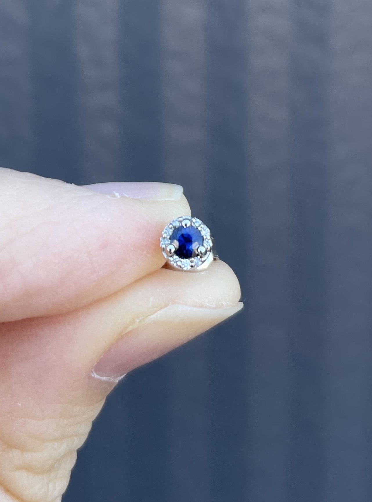 A close-up of a hand holding a small silver ring with a round blue sapphire center stone surrounded by a halo of Canadian diamonds, set against a dark gray textured background.
