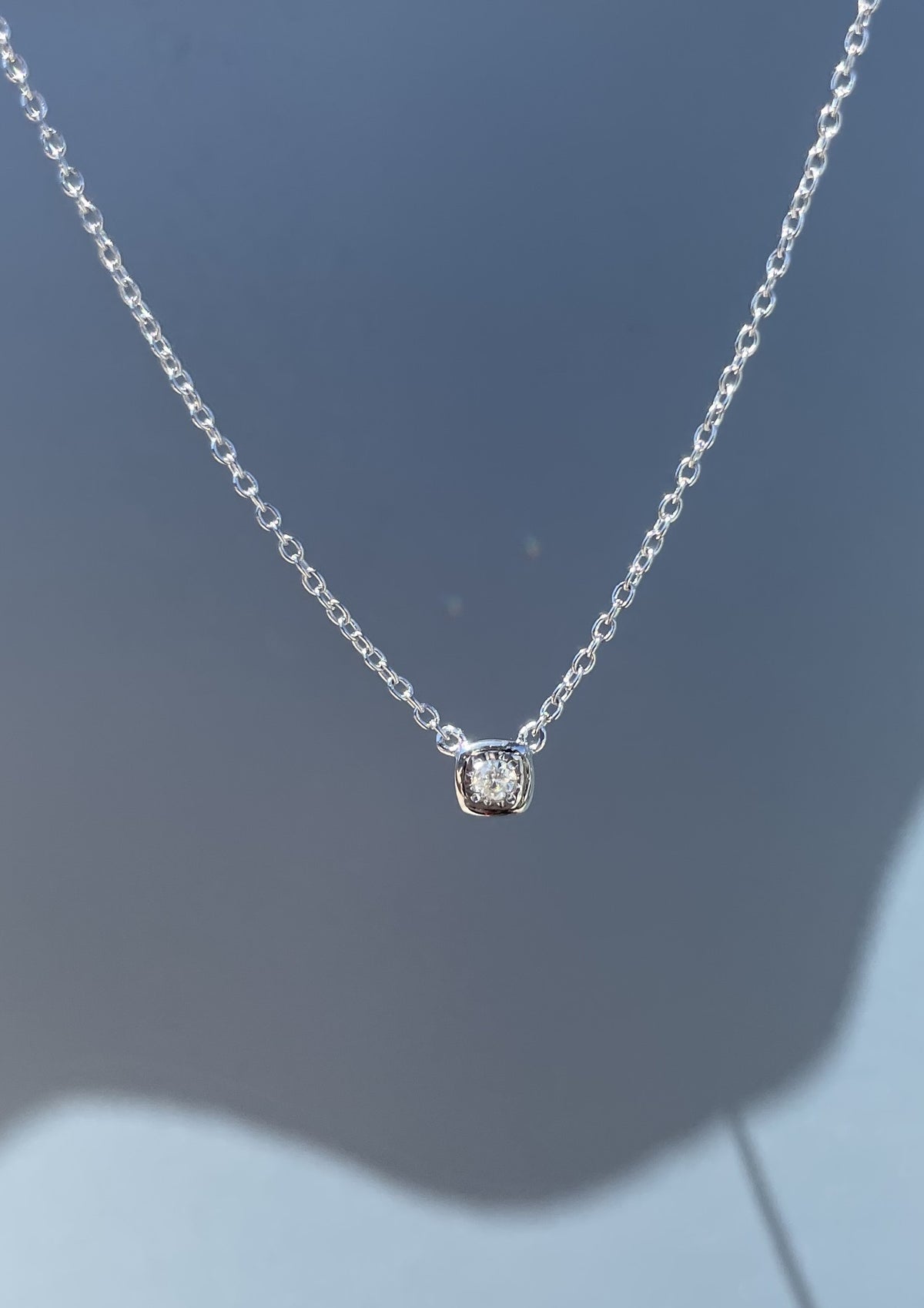 Close-up product view of a 925 sterling silver solitaire pendant necklace suspended against a grey background, showcasing a cushion-shaped miracle plate bezel setting that emphasizes the brilliance of the 0.04 Ct Canadian diamond focal stone.