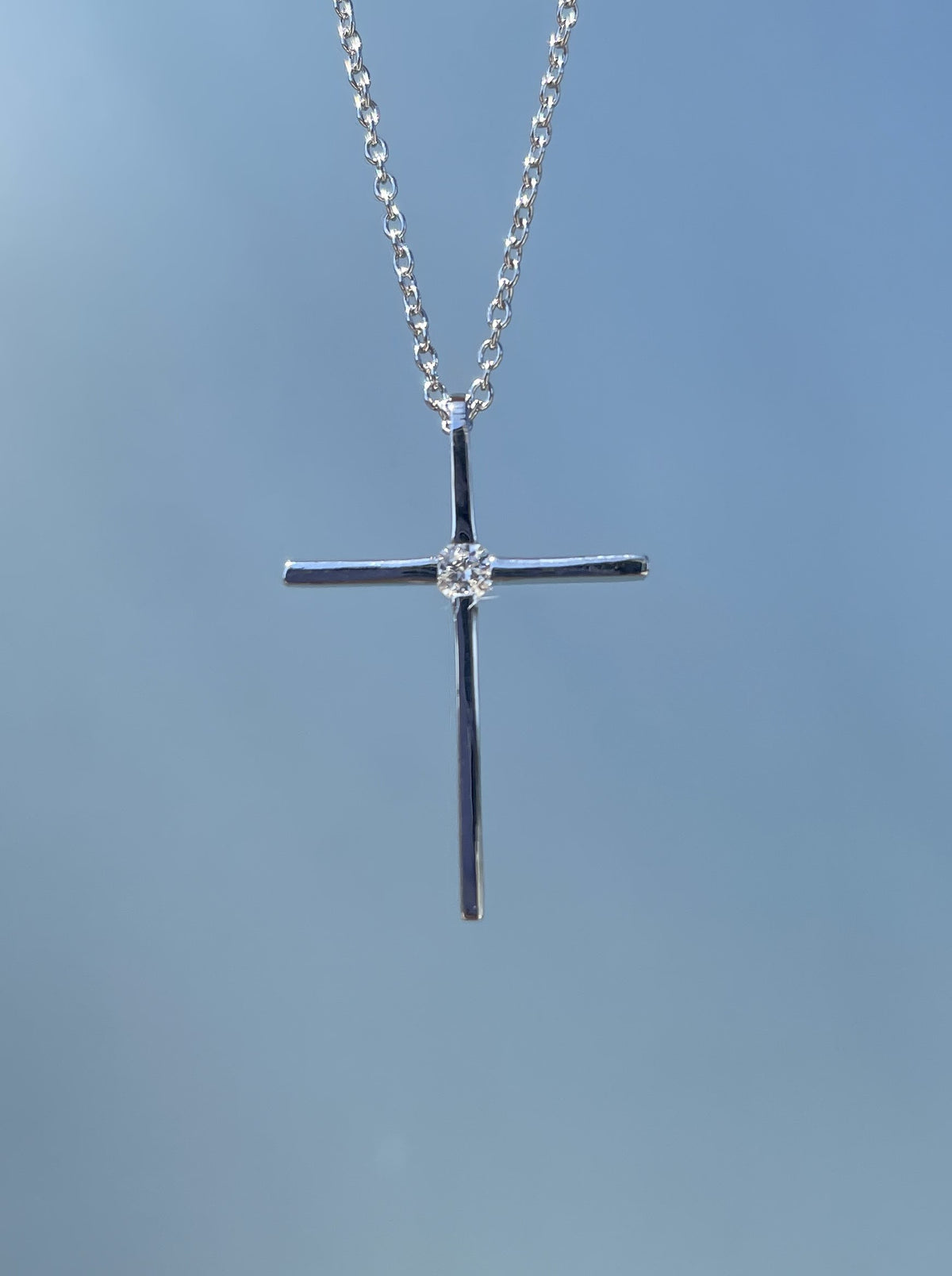 Close-up product view of a 925 sterling silver thin cross pendant necklace suspended against a grey-blue background, showcasing a highly polished minimalist frame accented by a single brilliant 0.12 Ct natural Canadian diamond solitaire at the center, displayed on a matching rolo chain.