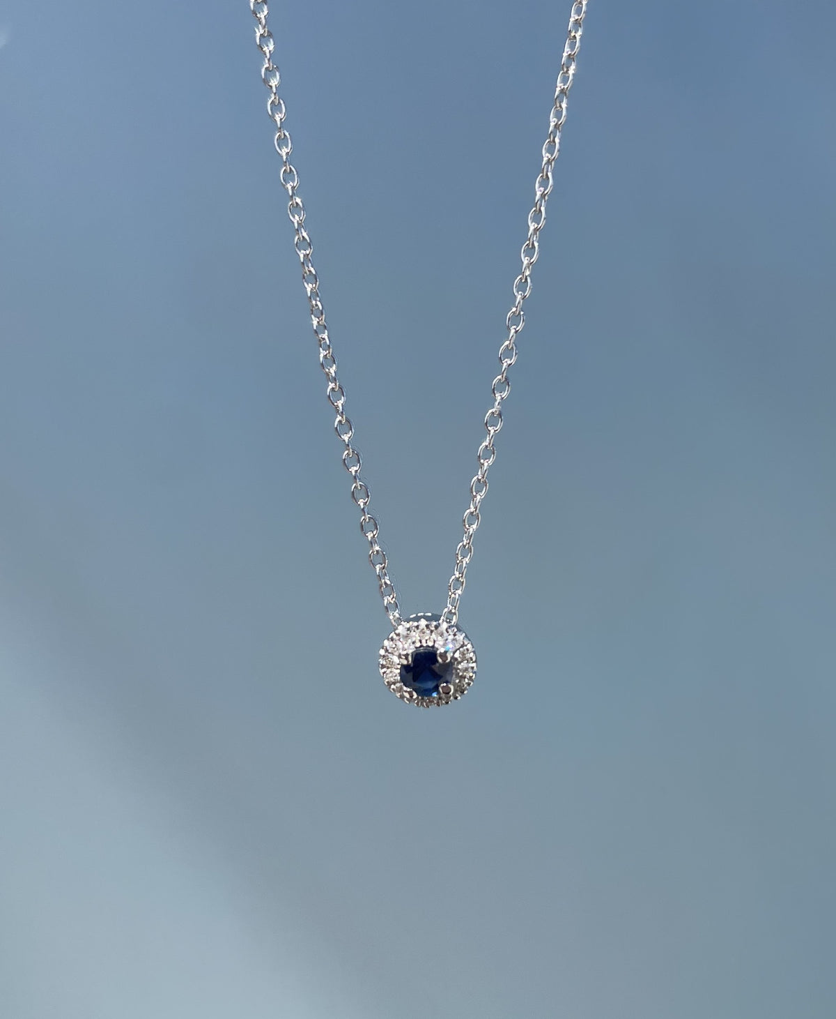Close-up product view of a 925 sterling silver halo pendant necklace suspended against a solid grey background, highlighting the round 0.18 Carat blue sapphire center stone closely encircled by a 0.10 Cttw Canadian diamond halo.