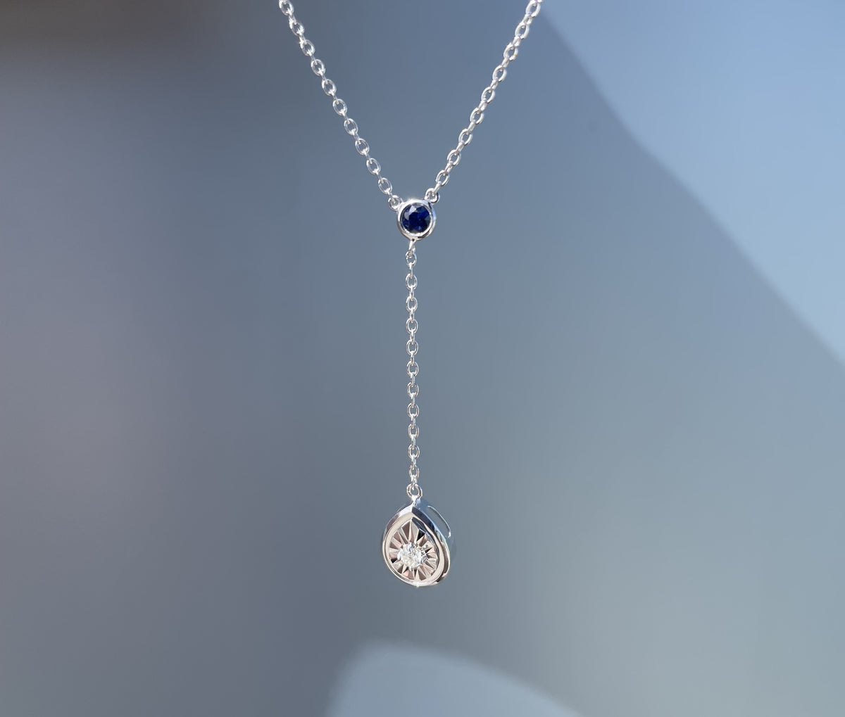 Close-up product view of a sterling silver Y-style pendant necklace suspended against a grey background, showcasing a round bezel-set blue sapphire at the chain junction and a dangling pear-shaped pendant featuring a miracle-set white diamond.