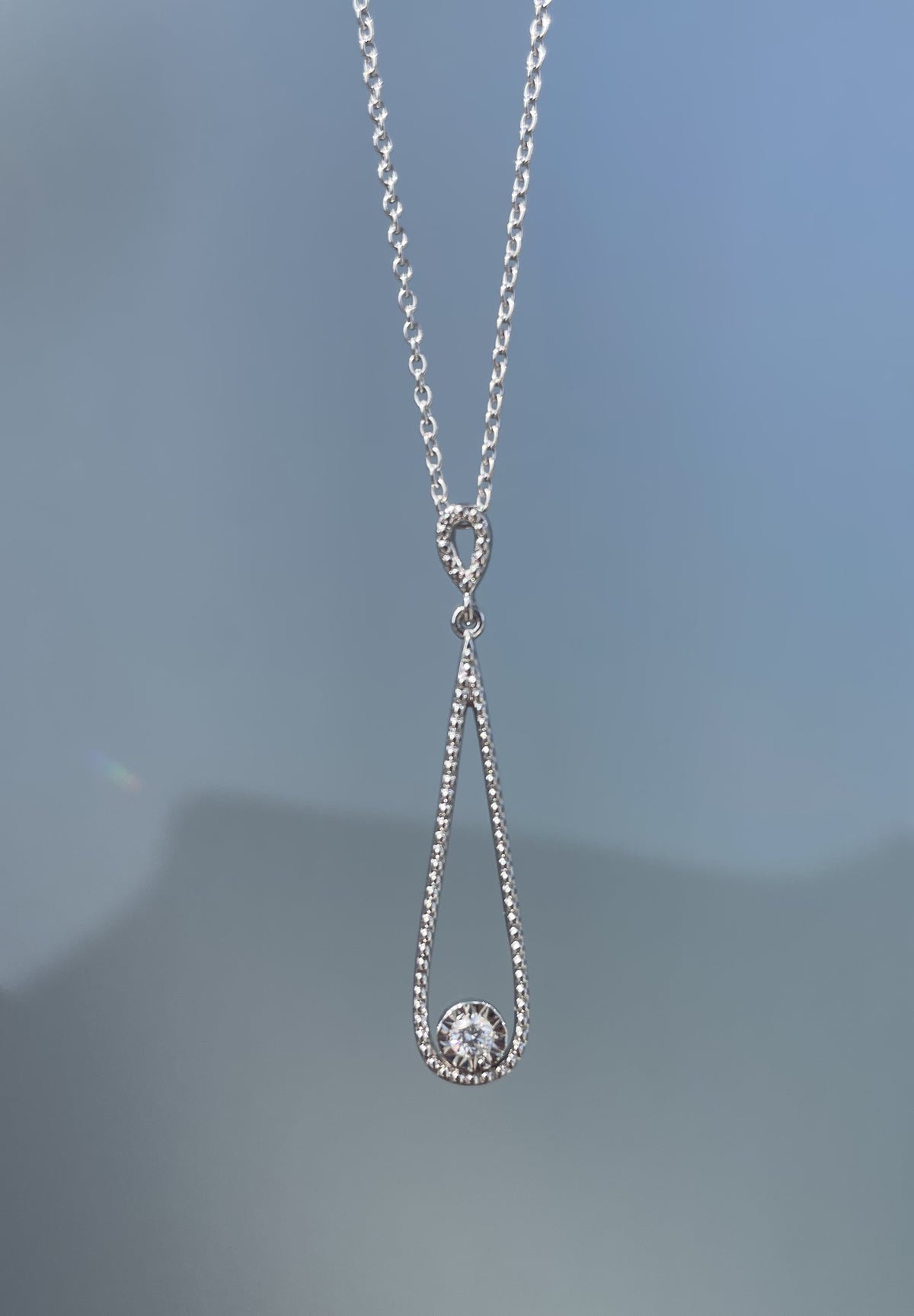 Close-up product view of a 925 sterling silver open teardrop pendant necklace suspended against a grey background, showcasing a textured milgrain style frame with a single 0.06 Ct round brilliant cut solitaire Canadian diamond nestled at the base.