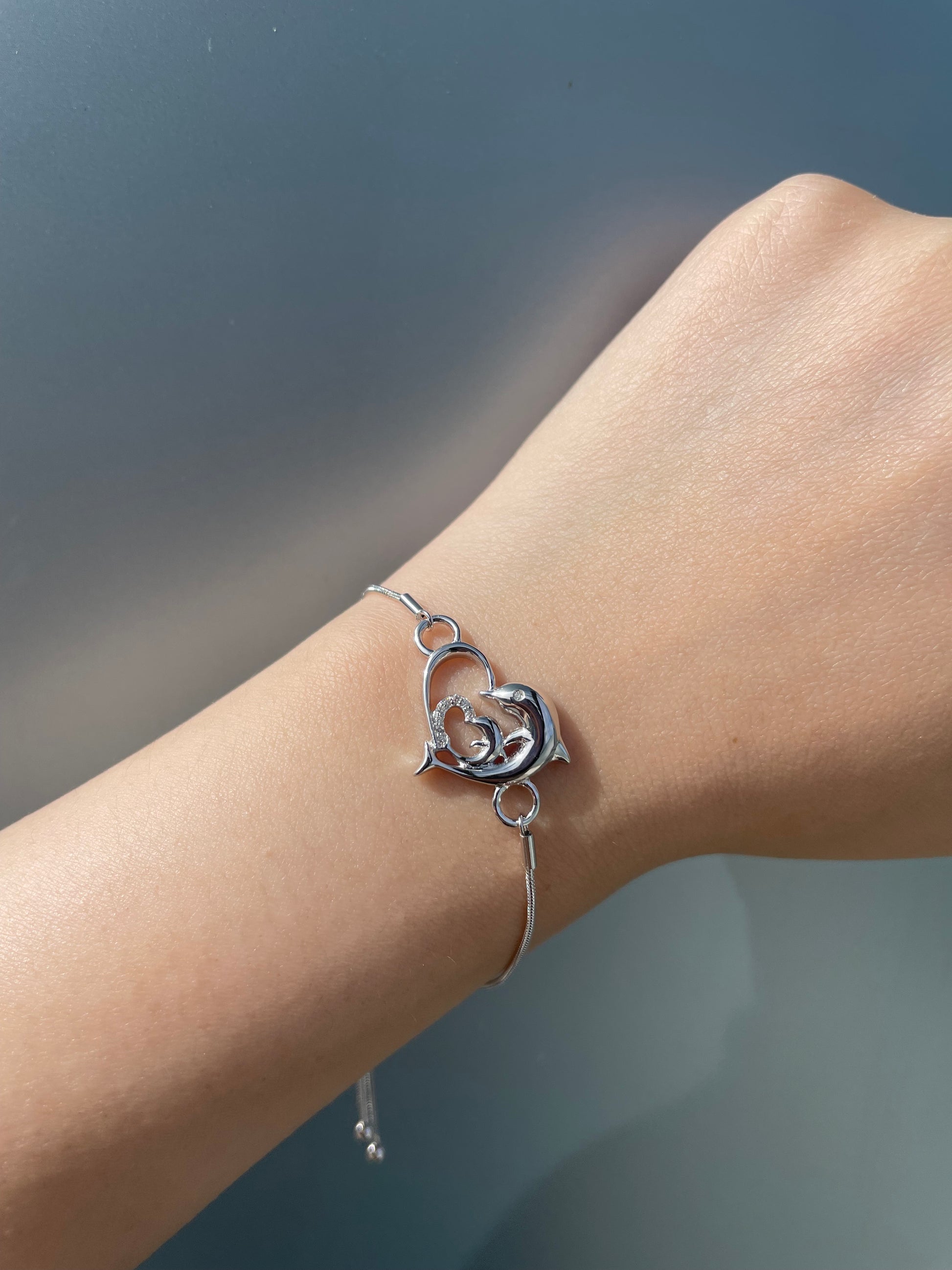 Close-up view of a 925 sterling silver bolo bracelet on a wrist against a solid grey background, highlighting the central open heart charm intertwined with dolphins design and 0.05 Cttw Canadian diamonds.