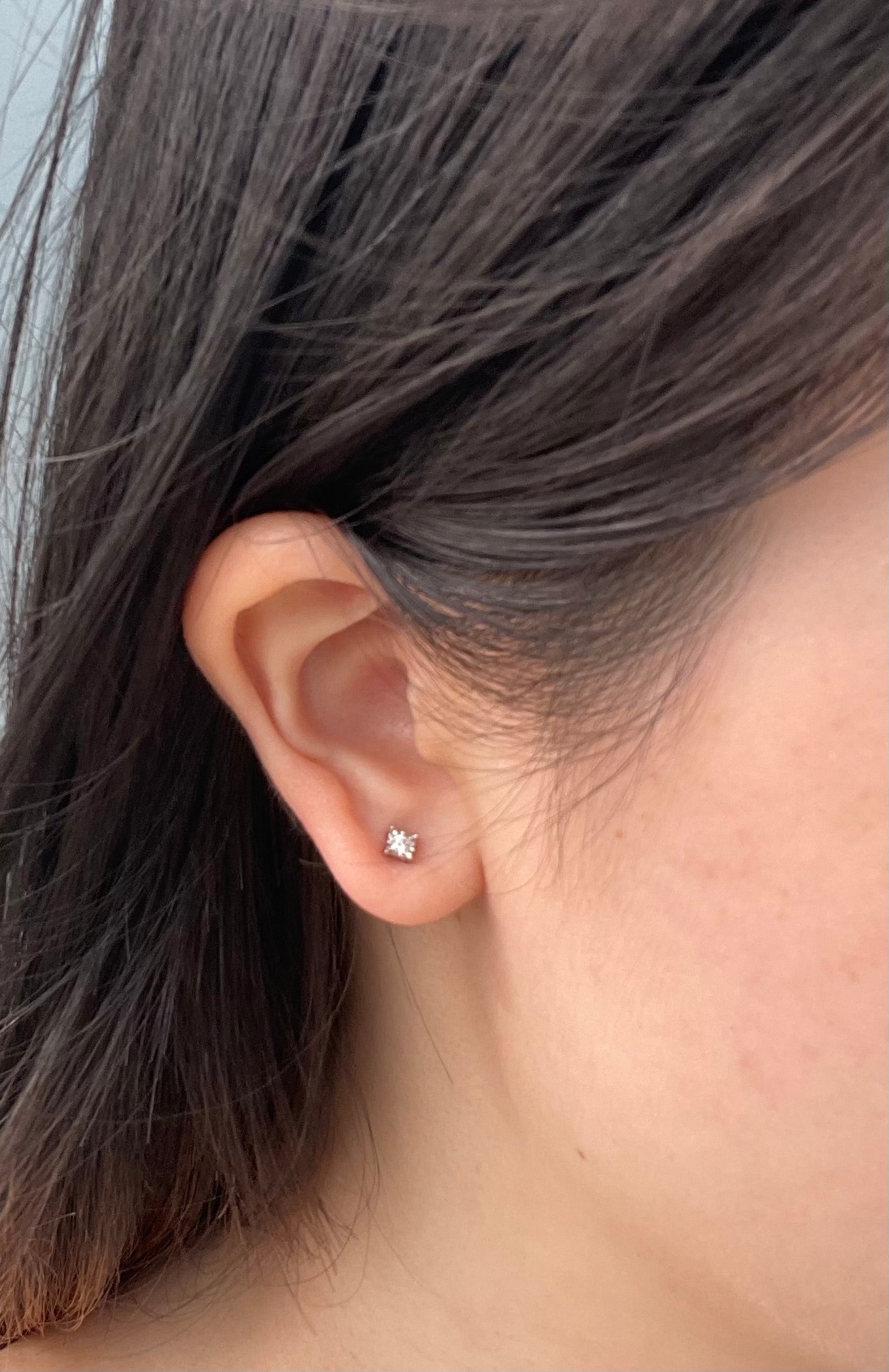 A model is wearing a single 925 Sterling Silver solitaire stud earring in her earlobe. The earring features a 0.03 carat Canadian Diamond secured in a Miracle-Setting, which is designed to enhance the stone's perceived size and sparkle. The close-up shot demonstrates the scale and appearance of the stud when worn.