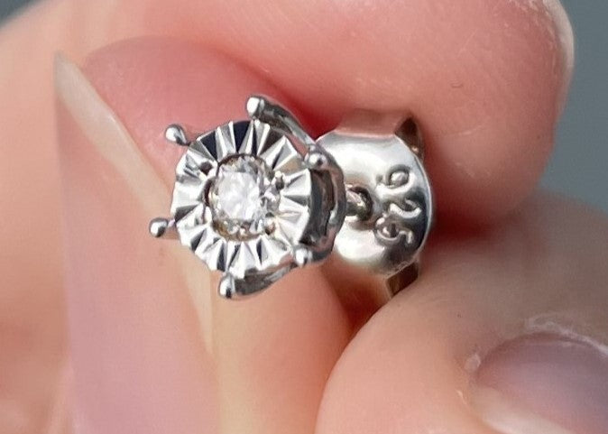 Extreme close-up product view of a 925 sterling silver solitaire stud earring held between fingers, highlighting the reflective miracle-setting and traditional 6-prong frame that enhances the brilliance of the 0.05 Ct Canadian diamond center stone, with the 925 metal hallmark visible on the butterfly earring back.