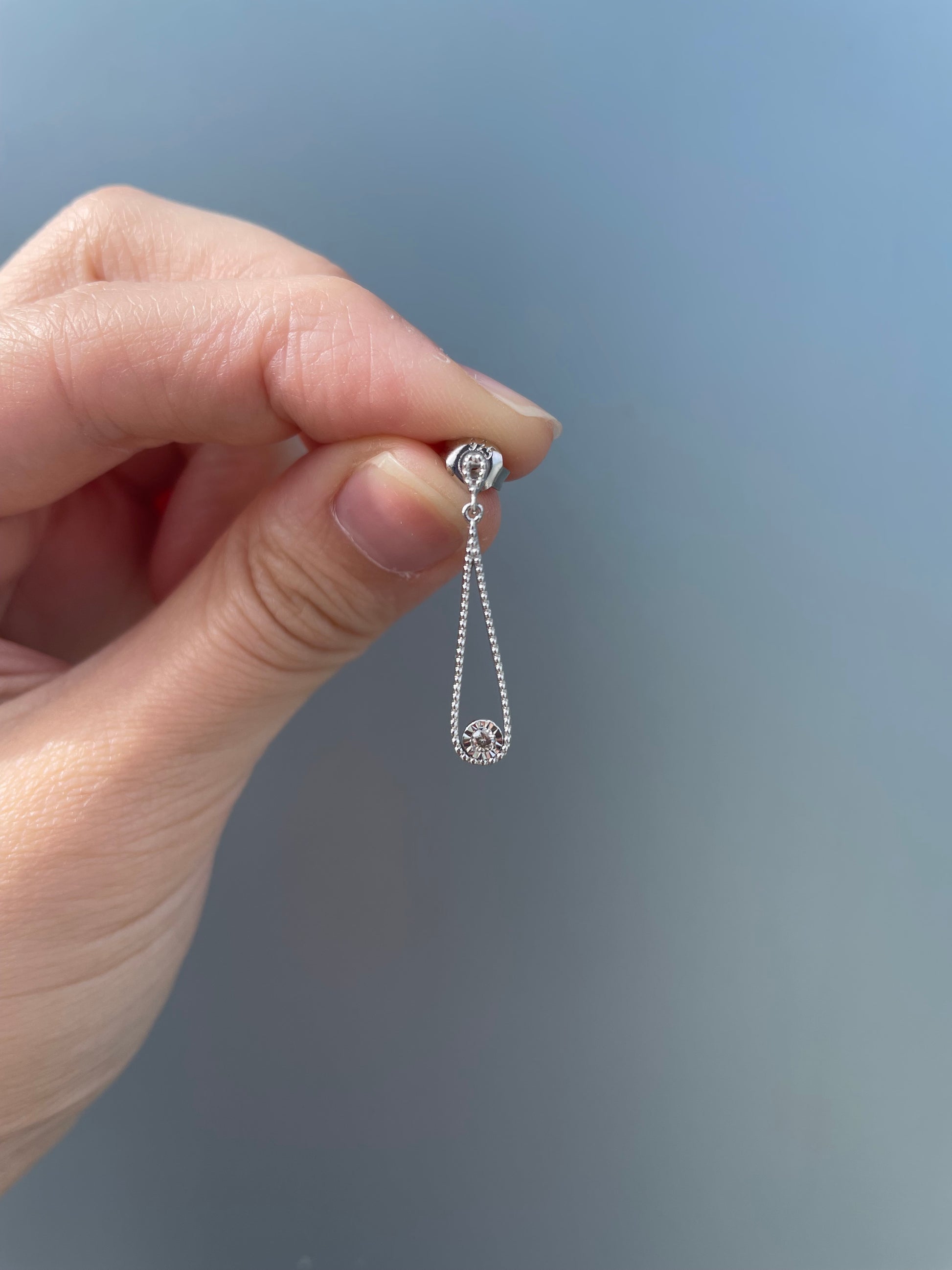 A hand holding a 925 sterling silver droplet dangle earring against a grey background, showcasing the open teardrop design with a beaded milgrain edge and a miracle-set Canadian diamond at the center base.