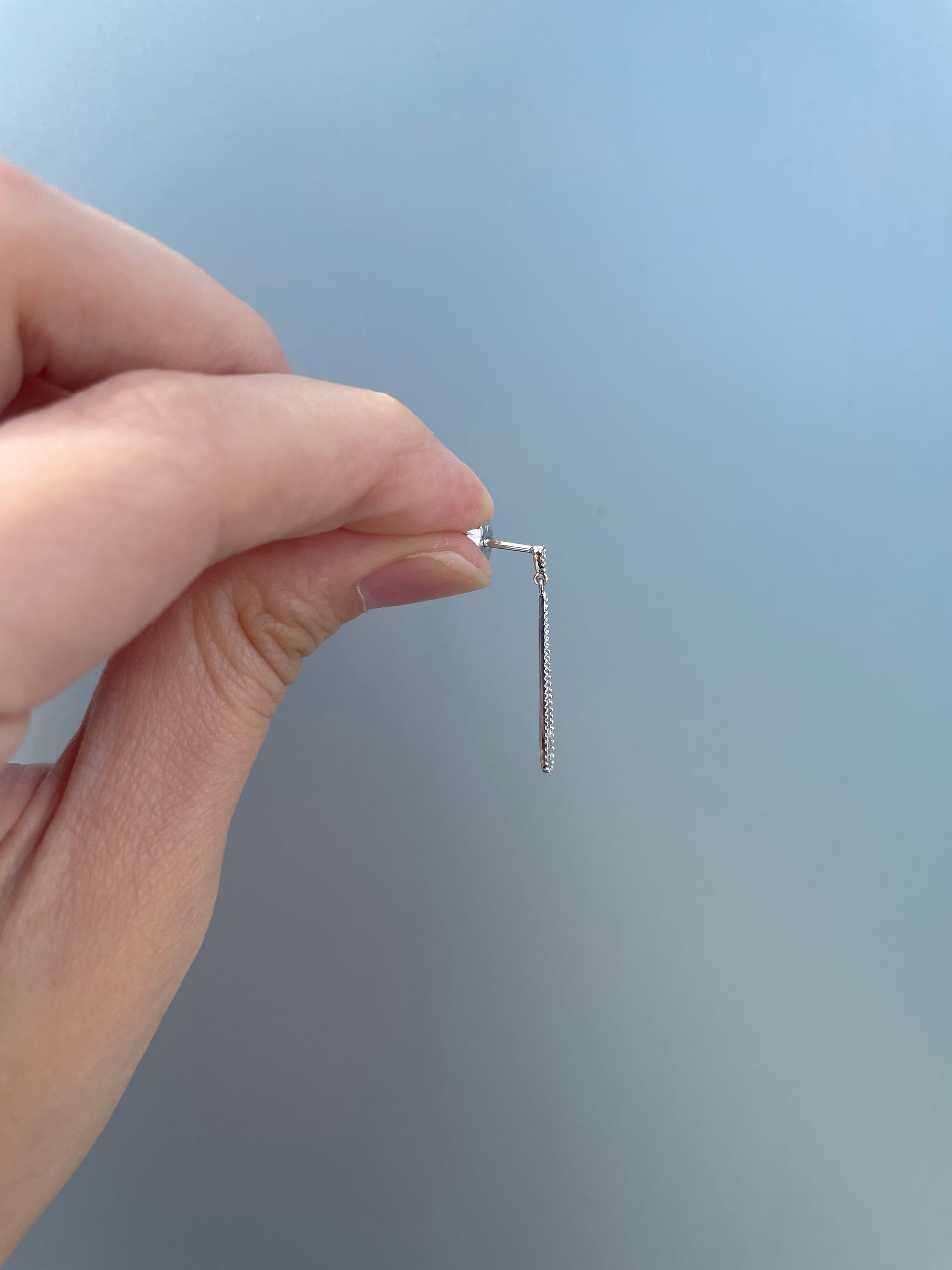 Side view profile of a 925 sterling silver droplet dangle earring held against a grey background, showcasing its slim profile, the earring post construction, and the delicate milgrain design edge.