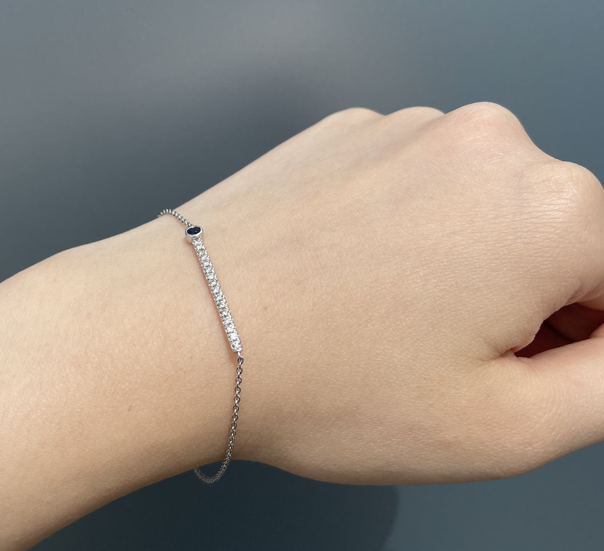 A delicate silver chain bracelet with a row of Canadian diamonds and a blue sapphire accent, shown worn on a person's wrist against a dark gray background.