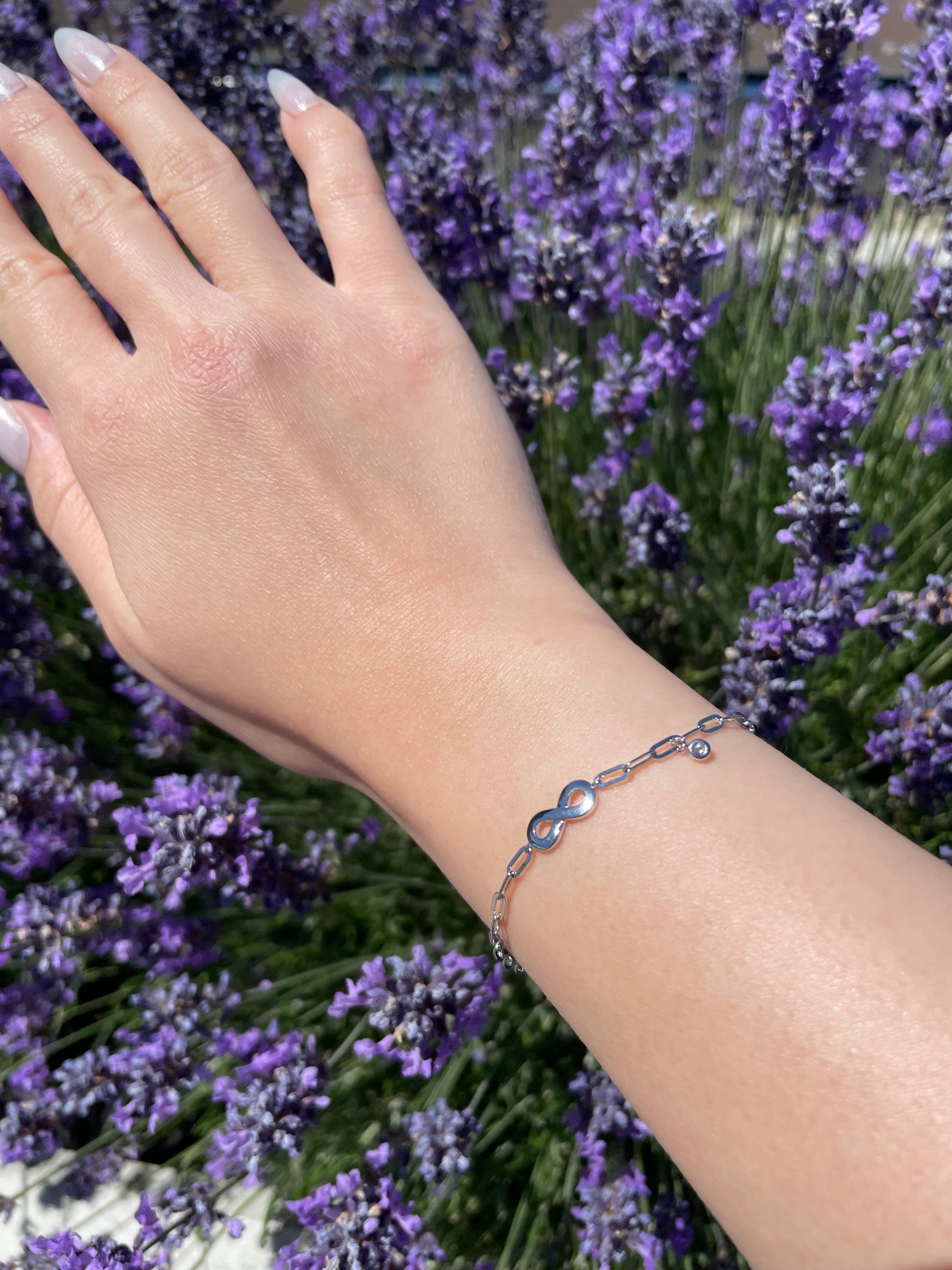 Close-up of a model's wrist wearing a sterling silver paperclip chain bracelet featuring a polished infinity symbol center station and a delicate bezel-set diamond charm, set against a vibrant background of blooming purple lavender.