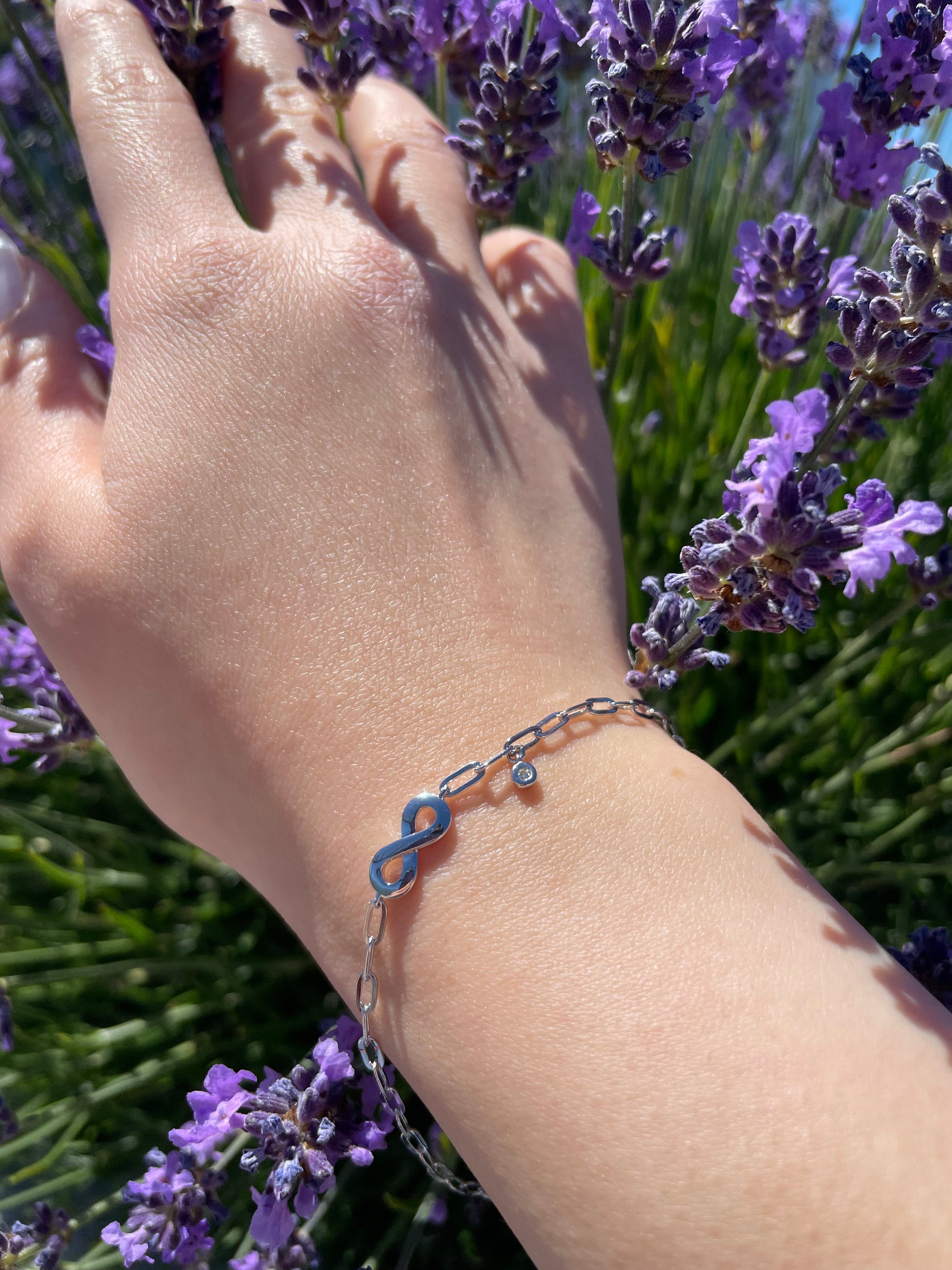 Close-up of a model's wrist wearing a sterling silver paperclip chain bracelet featuring a polished infinity symbol center station and a delicate bezel-set diamond charm, set against a vibrant background of blooming purple lavender.