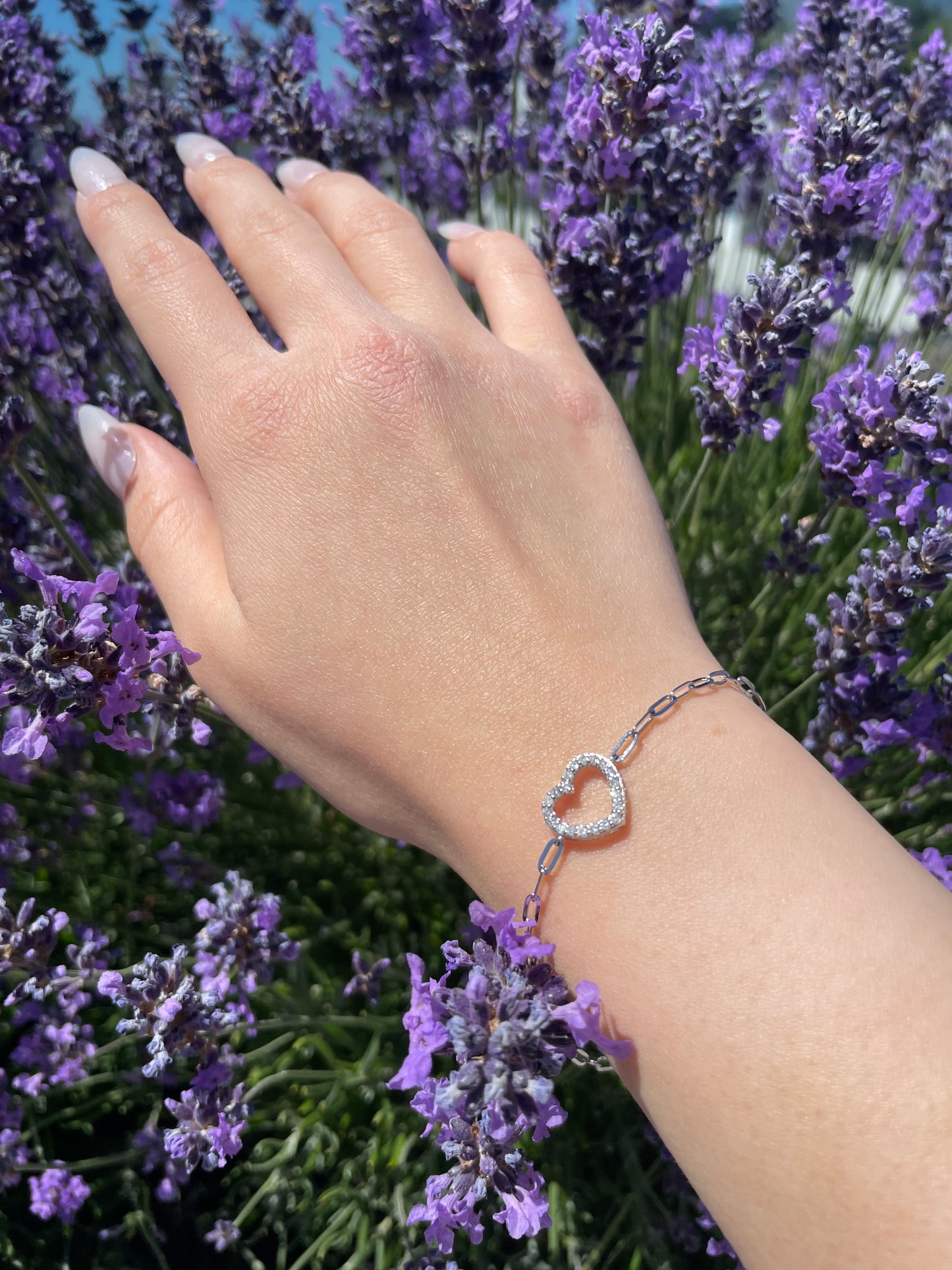 Close-up of a model's wrist wearing a silver paperclip chain bracelet featuring an open heart center with shimmering prong set Canadian diamonds, set against a backdrop of deep purple lavender.
