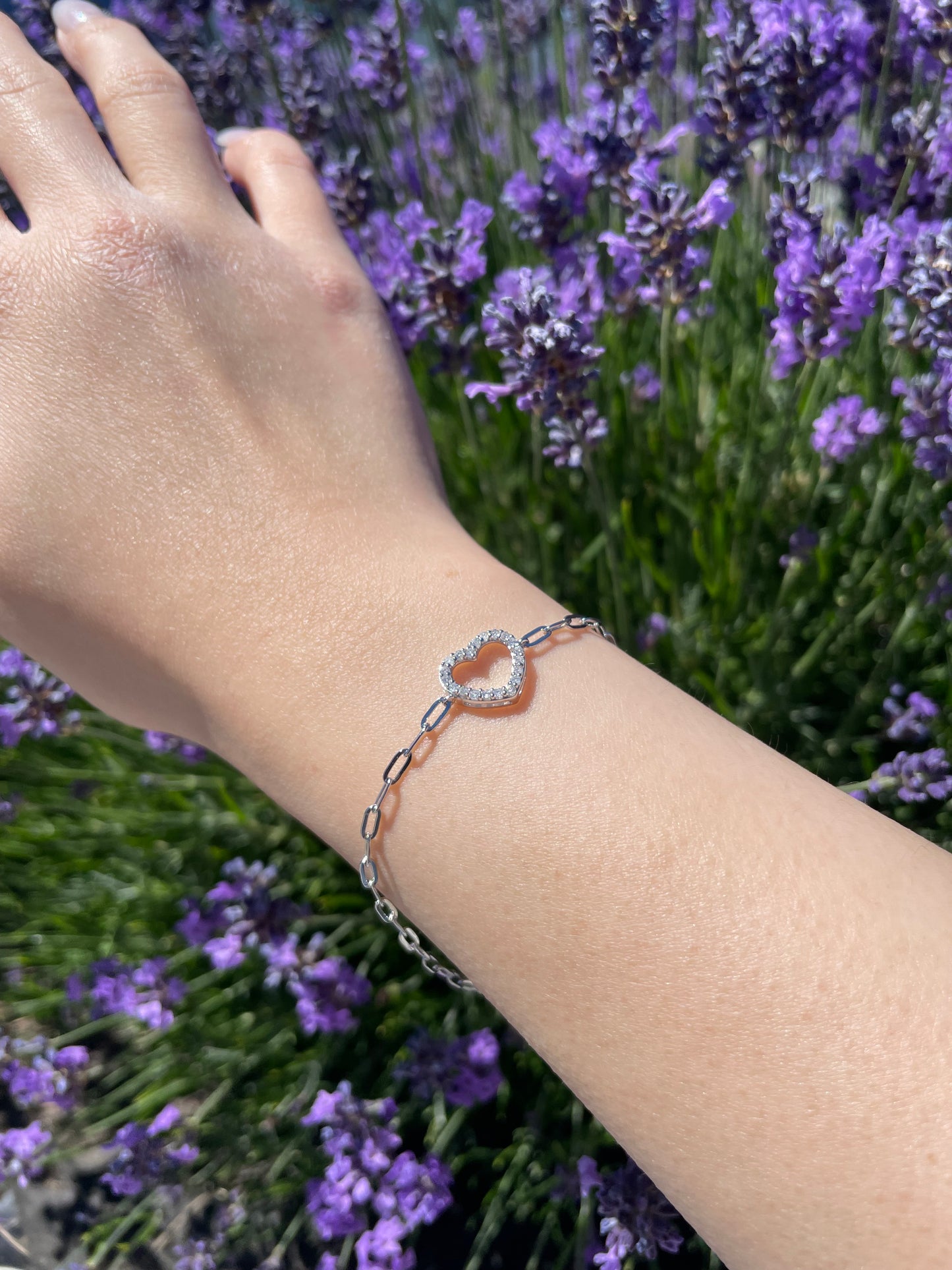 Lifestyle image of a silver paperclip chain bracelet showcasing a sparkling Canadian diamond open heart design worn on a wrist, captured in natural bright daylight with a blurred background of purple lavender flowers.