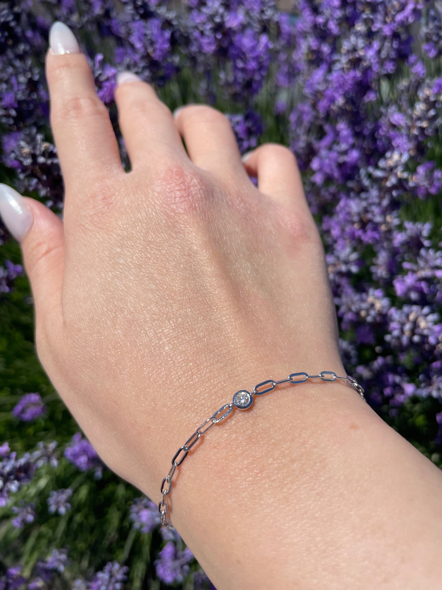 Close-up lifestyle shot of a model's wrist wearing a silver paperclip chain bracelet featuring a single, brilliant round bezel-set Canadian diamond at the center, showcased against a vibrant background of blooming purple lavender.