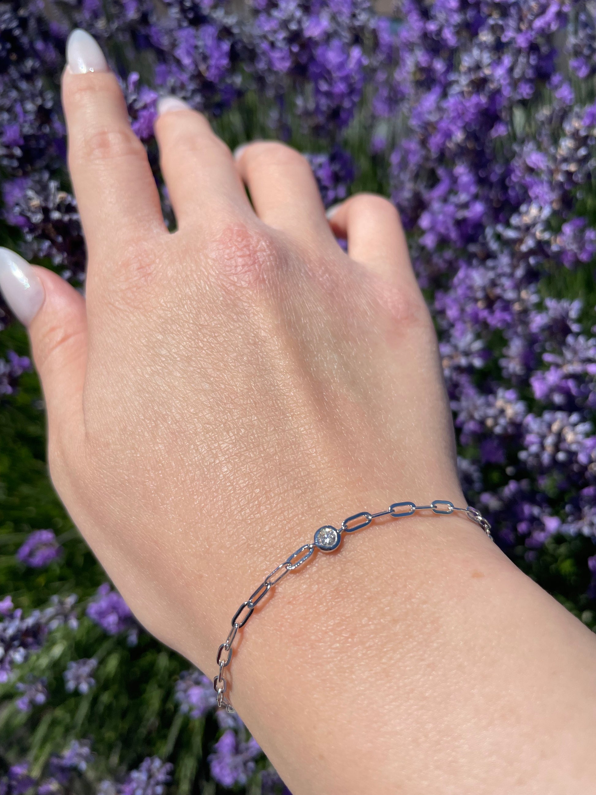 Close-up lifestyle shot of a model's wrist wearing a silver paperclip chain bracelet featuring a single, brilliant round bezel-set Canadian diamond at the center, showcased against a vibrant background of blooming purple lavender.