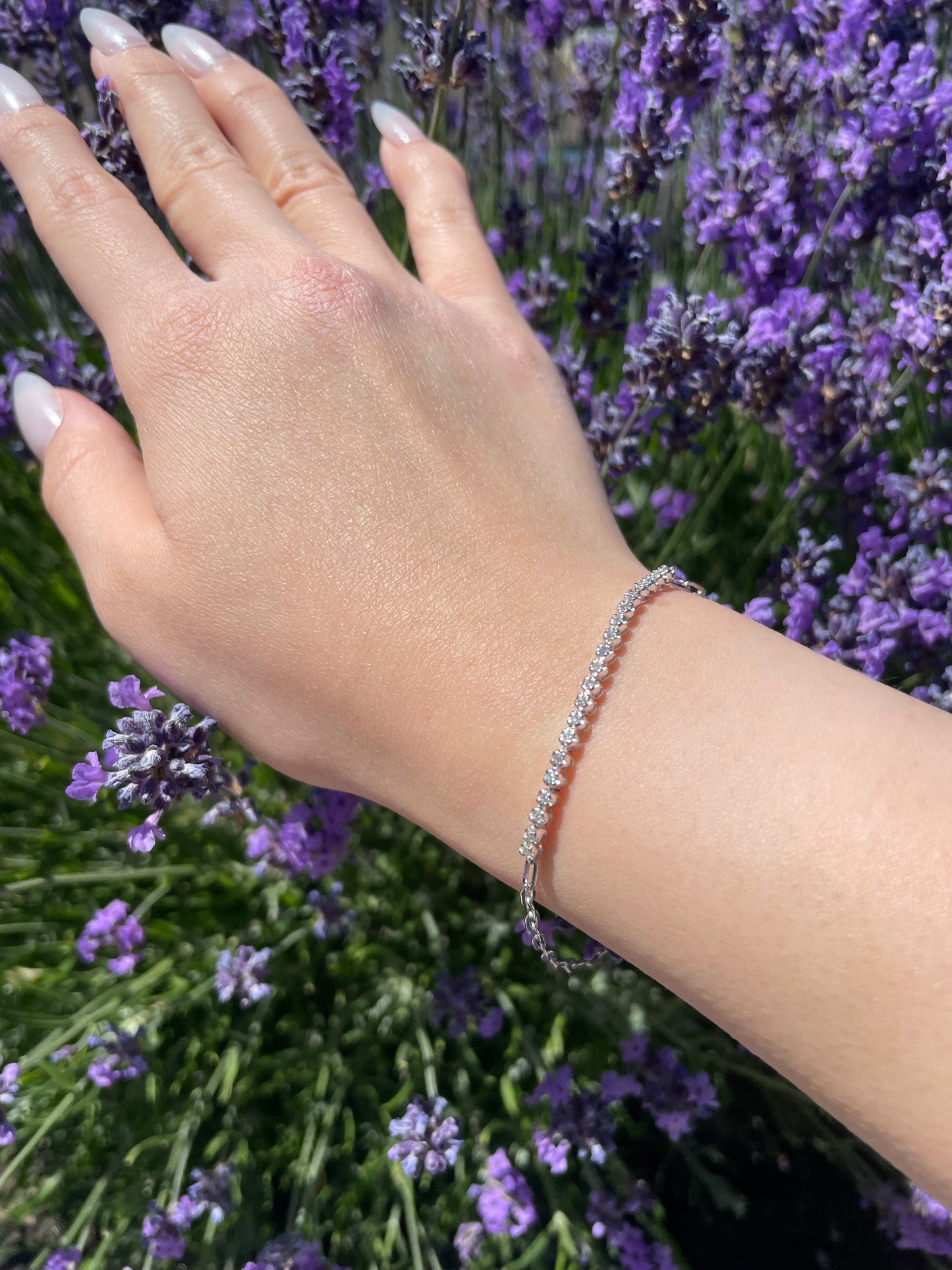 A close-up outdoor lifestyle shot of a model's wrist displaying a sterling silver half tennis paperclip link bracelet. The bracelet features a continuous, sparkling row of prong-set Canadian diamonds along the top of the wrist, transitioning into modern paperclip chain links. The model's hand rests gently against a vibrant, sunlit background of purple lavender stalks.