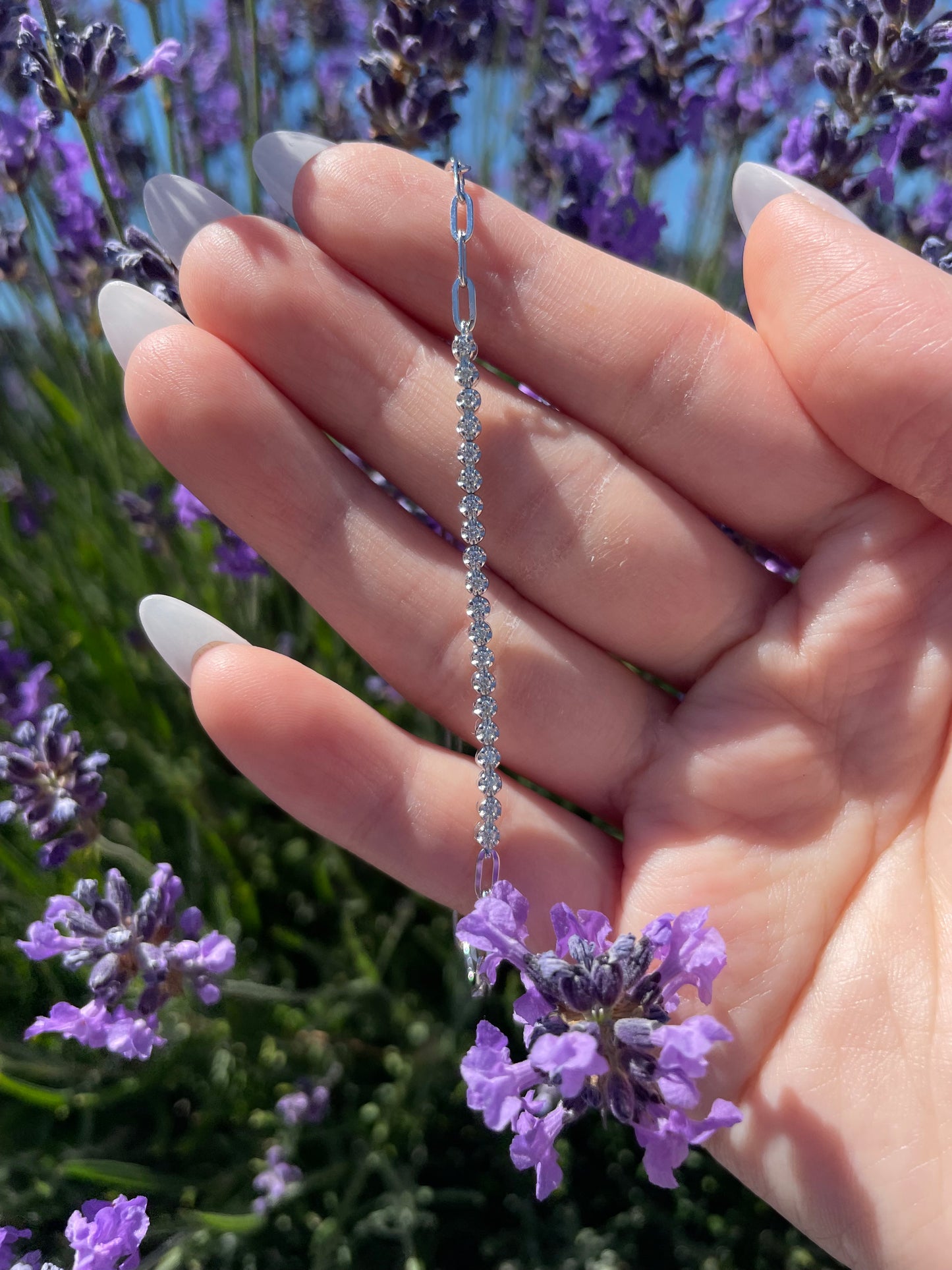 An outdoor lifestyle shot showing a delicate sterling silver 0.22 ctw Canadian diamond half tennis paperclip chain bracelet draped vertically across a model's open fingers. The design features a brilliant, continuous row of prong-set Canadian diamonds along the tennis section, transitioning into modern, high-polished paperclip links. The hand is held against a vibrant, sunlit background of purple lavender stalks, emphasizing the bracelet's clean shine and fine scale.