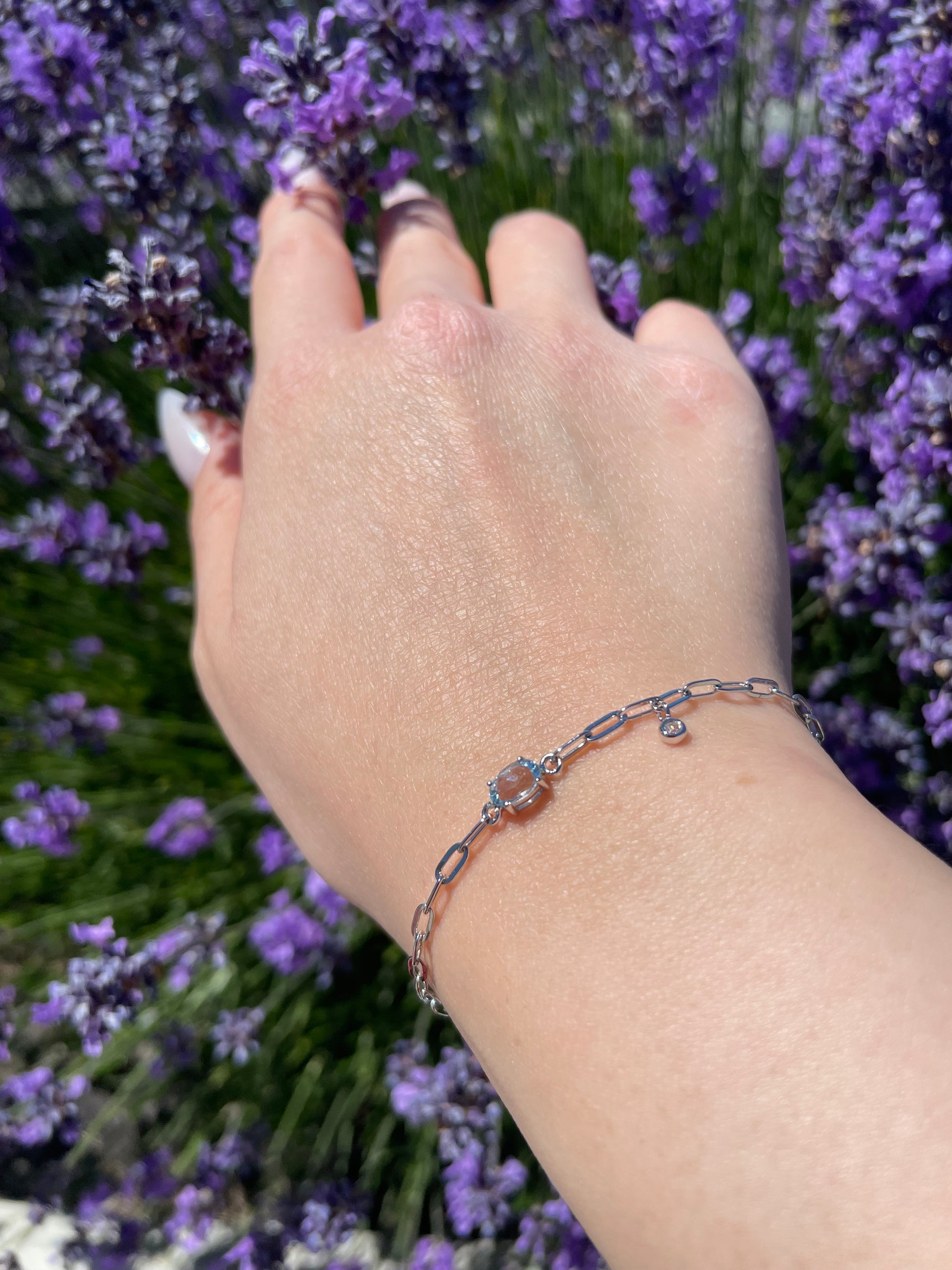 Hand wearing a 925 Sterling Silver 6x4MM Natural Aquamarine and Canadian Diamond Paperclip Bracelet with lavender flowers in the background