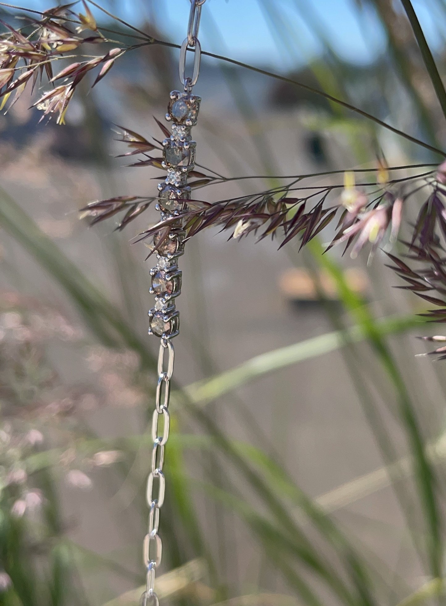 A vertical close-up shot of a silver paperclip chain bracelet hanging among tall field grass. The center station features an alternating pattern of 6 prong-set round Canadian coffee diamonds and 5 brilliant white Canadian diamonds, showcased in natural light.