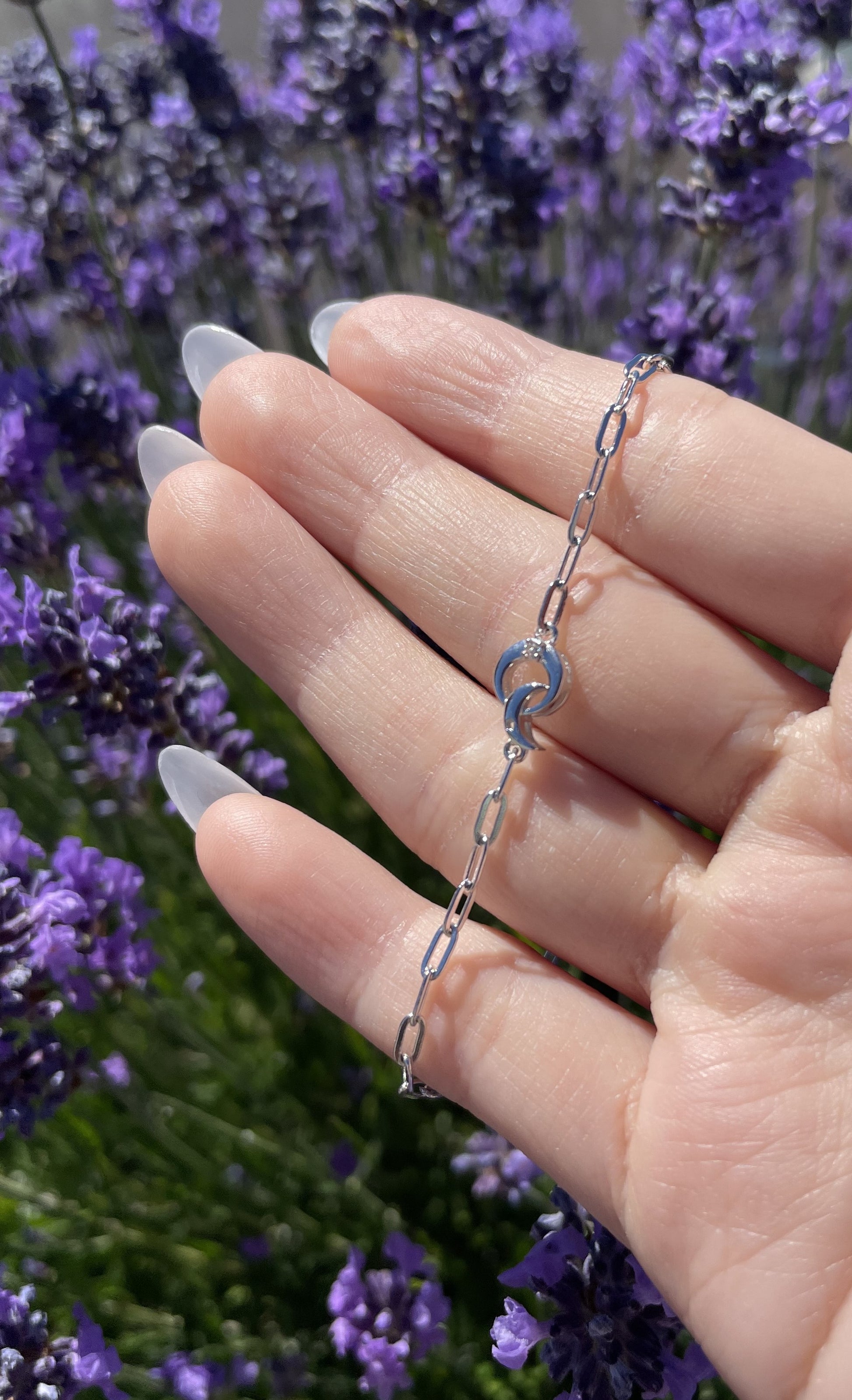 An outdoor lifestyle shot showing a delicate sterling silver paperclip chain bracelet laid across a model's open fingers. The central charm features a modern interlocking design of a textured crescent moon and a polished open circle, accented with a single sparkling Canadian diamond. The hand is set against a vibrant, brightly sunlit background of blooming purple lavender flowers, emphasizing the jewelry's clean metallic shine and petite scale.
