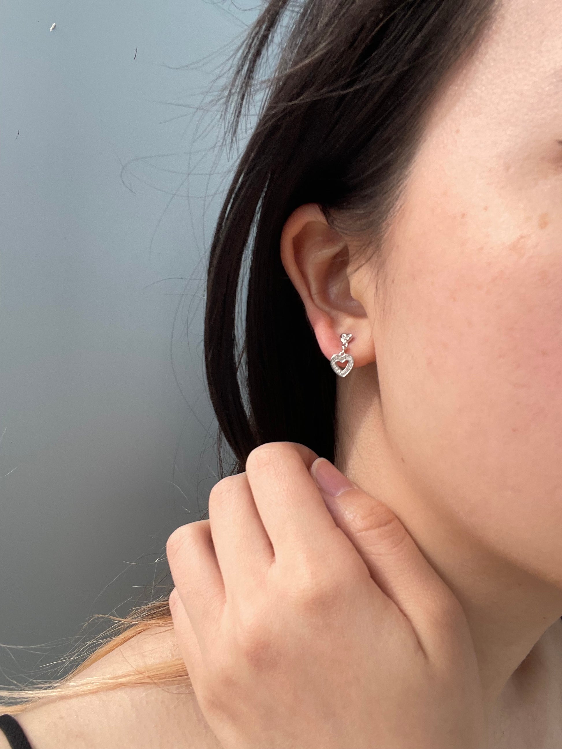 Close-up model photo showcasing a 925 sterling silver dangle earring on an earlobe, highlighting the small post stud that drops into an open heart charm paved with 0.10 Cttw Canadian diamonds.