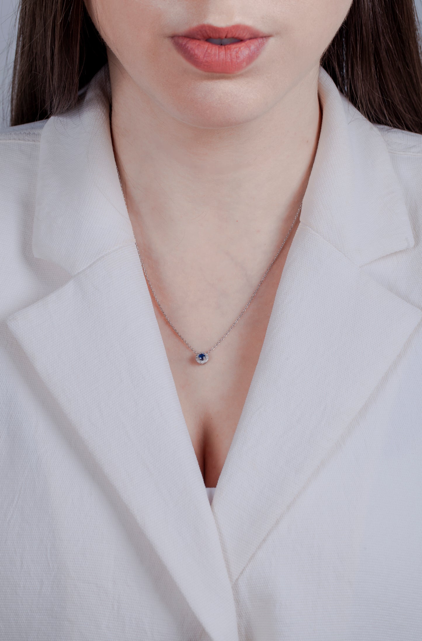 Person with brown long hair in a white blazer wearing a sterling silver sapphire and diamond halo necklace against a neutral background