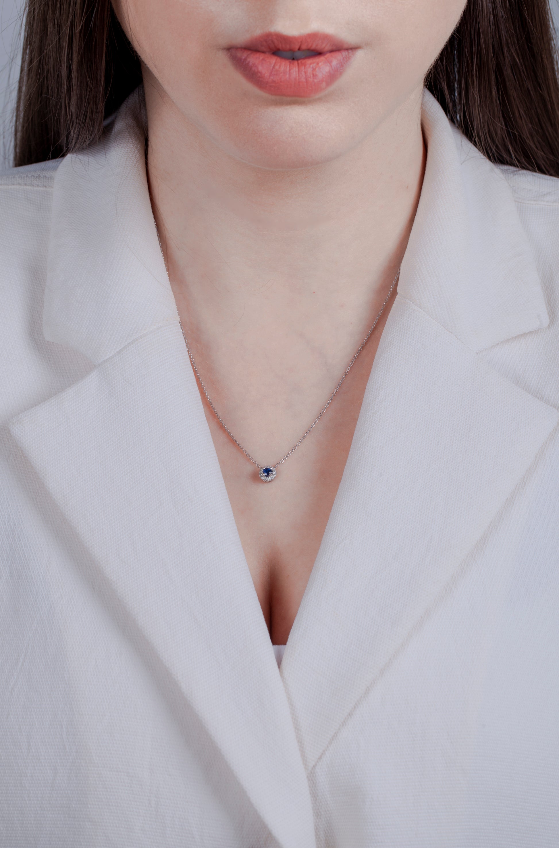 Person with brown long hair in a white blazer wearing a sterling silver sapphire and diamond halo necklace against a neutral background