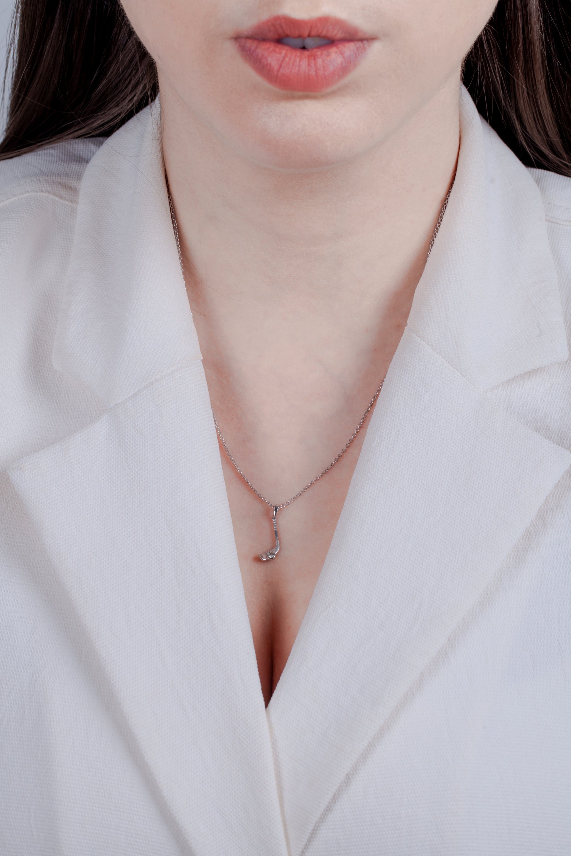 Model in a white blazer wearing a matte finish solid 925 sterling silver hockey stick pendant. A 0.05 carat Canadian diamond is set at the base of the blade, designed to look like a hockey puck. This matte, minimalistic charm hangs from an 18-inch rolo chain.