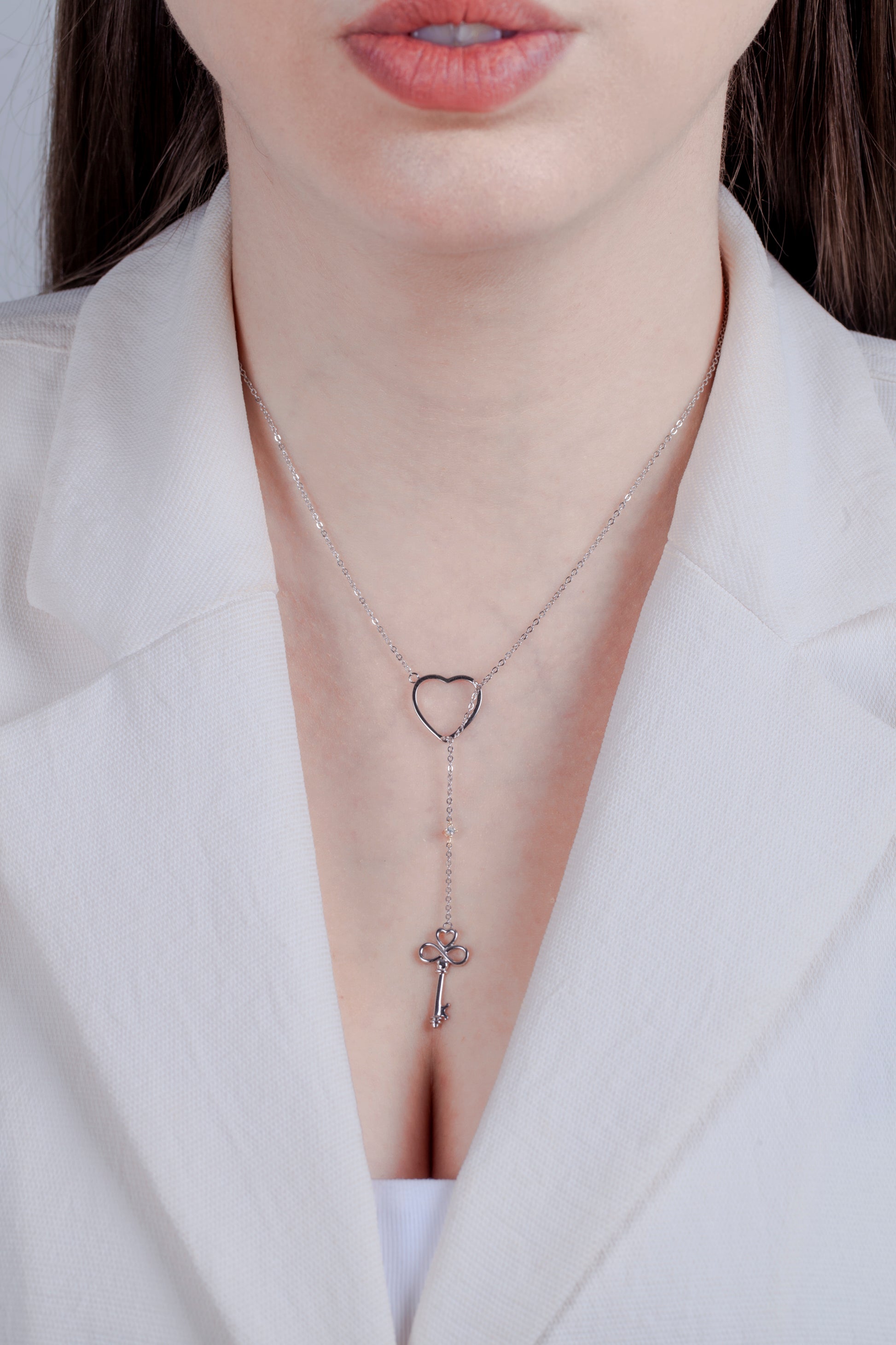 Close-up of a model wearing a white blazer, styling the sterling silver 'Key to My Heart' lariat necklace. The Y-shape of the drop necklace is highlighted against her skin, showing the scale of the heart slider, the dainty diamond accent, and the decorative key pendant.