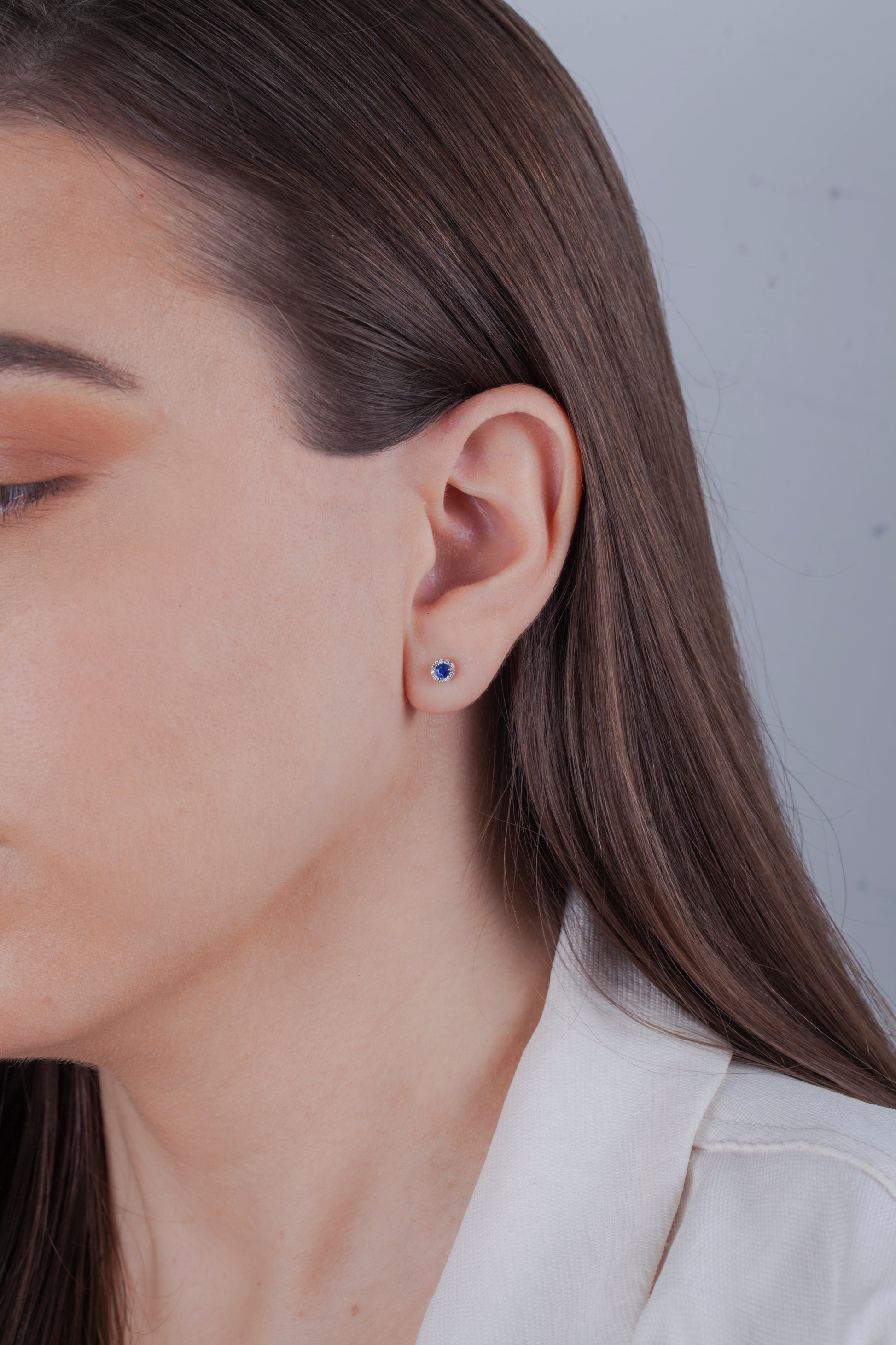 Close-up of a person wearing a small earring with a sapphire and Canadian diamond halo, brown long hair and white blazer against a neutral background