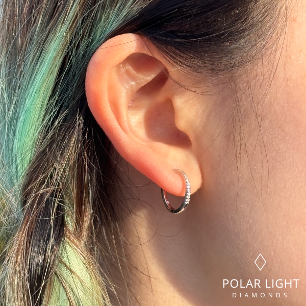 A close-up side profile of a model with vibrant dyed green and brown hair wearing a 925 sterling silver huggie hoop earring. The small hoop is lined with a row of sparkling Canadian diamonds that catch the sunlight, highlighting the contrast between the polished silver and the colorful hair. The Polar Light Diamonds logo is visible in the bottom right corner.